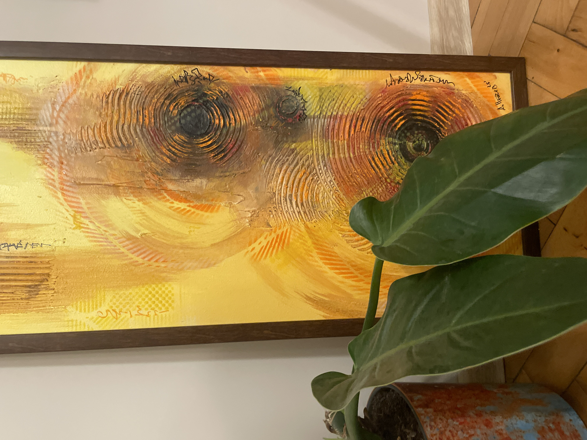 A framed abstract painting with dominant yellow and orange hues hangs on a plain wall. The painting features concentric circles and textured lines, with dark centers within some of the circles. A signature and what appears to be a date, "25'", are visible in the lower right corner. In the foreground, a leafy green plant in a mottled orange and blue pot partially obscures the bottom left of the painting. The flooring is wooden planks. The scene is indoors and appears to be a home setting. There are no people or animals visible.