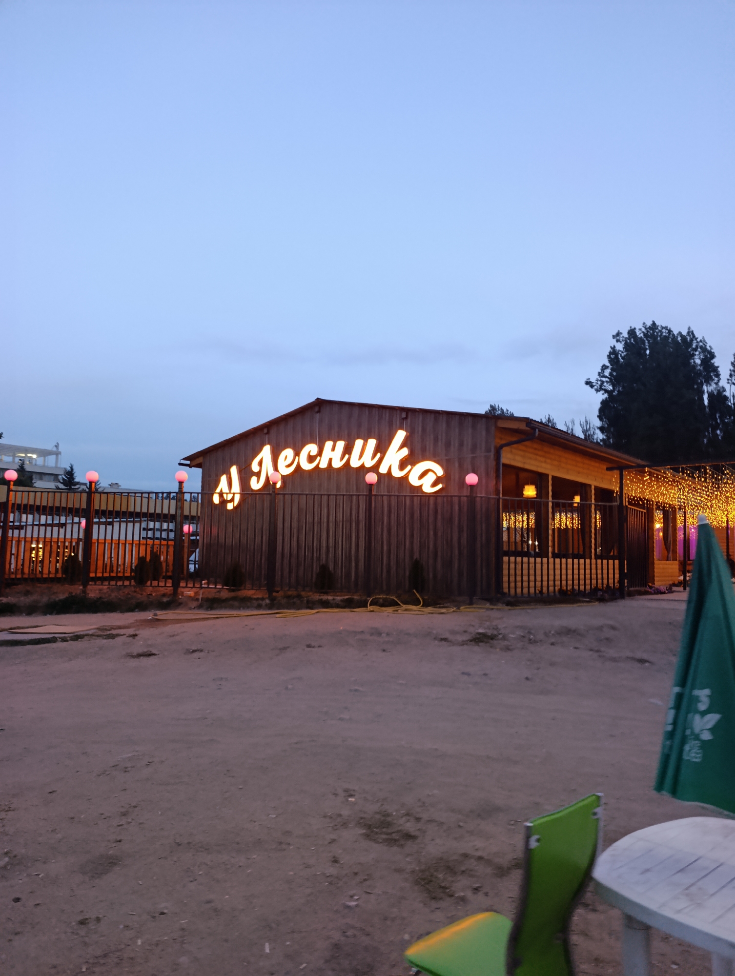 The image depicts an outdoor scene at dusk, likely a restaurant or cafe, with the signage "ЛЕСНИКА" illuminated in neon on a wooden building. The building features wooden siding and a covered outdoor area adorned with strings of warm-toned lights. A metal fence runs along the front of the property, with decorative pink spherical lights mounted on posts. In the foreground, there is a dirt ground surface, a green patio umbrella, a white circular table, and a green chair. The background shows trees and some distant structures. The overall atmosphere is calm and inviting, suitable for an evening gathering. No people are visible in the image.