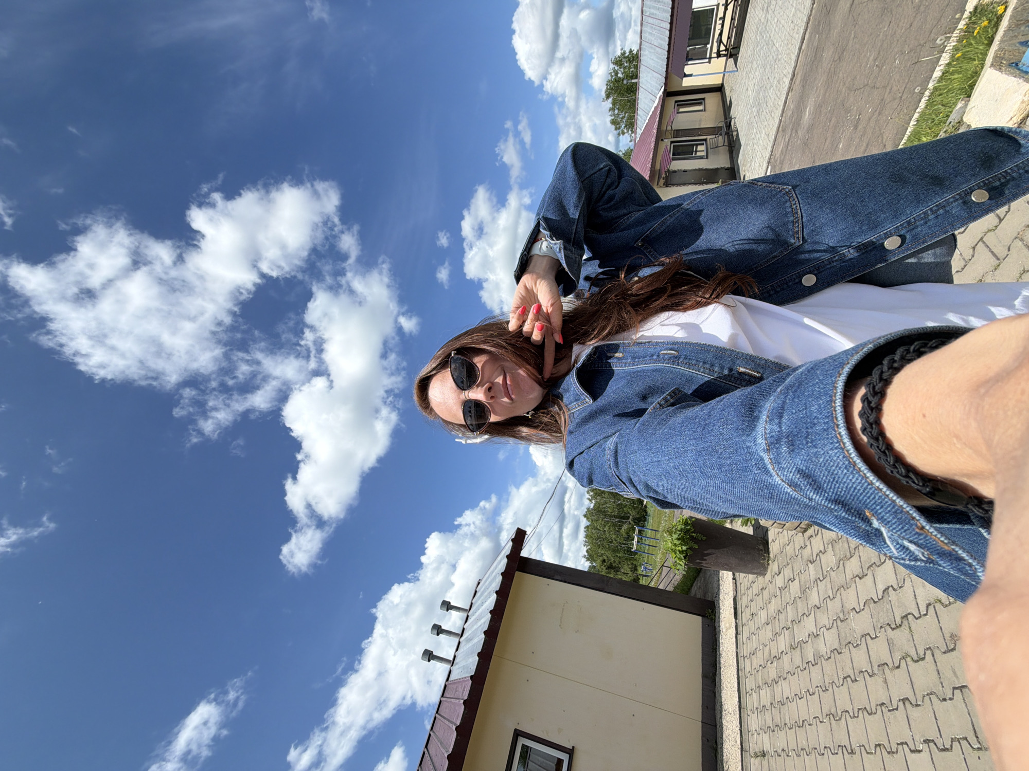 A woman with long brown hair, wearing a denim jacket, sunglasses, and a white shirt, poses for a selfie outdoors on a sunny day with a blue sky and white clouds. She is standing in front of a yellow building with a red-brown roof, which appears to be part of a small complex. The ground is paved with interlocking bricks. In the background, there are trees and other buildings. The overall scene suggests a casual outdoor setting, possibly a rural or suburban area. The image has a wide-angle distortion due to the selfie perspective. Without further information or context, it is impossible to definitively determine if this location is in Birobidzhan, Russia.