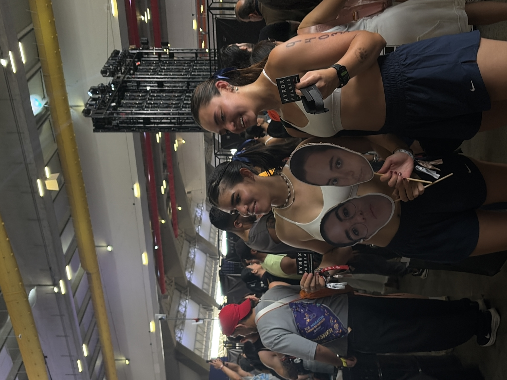 Two young women are the focal point of the image. The woman on the right is holding a black rectangular card with white text "HYROX PUEBLA", possibly a promotional or event-related item. The woman on the left is holding up two cardboard masks of individuals. Both women are wearing activewear, suggesting they might be involved in a physical activity or event. They are in an indoor setting, possibly an event venue, evidenced by the complex overhead lighting system and other people in the background. The lighting includes prominent yellow support beams, with a dark and busy background with lots of people.