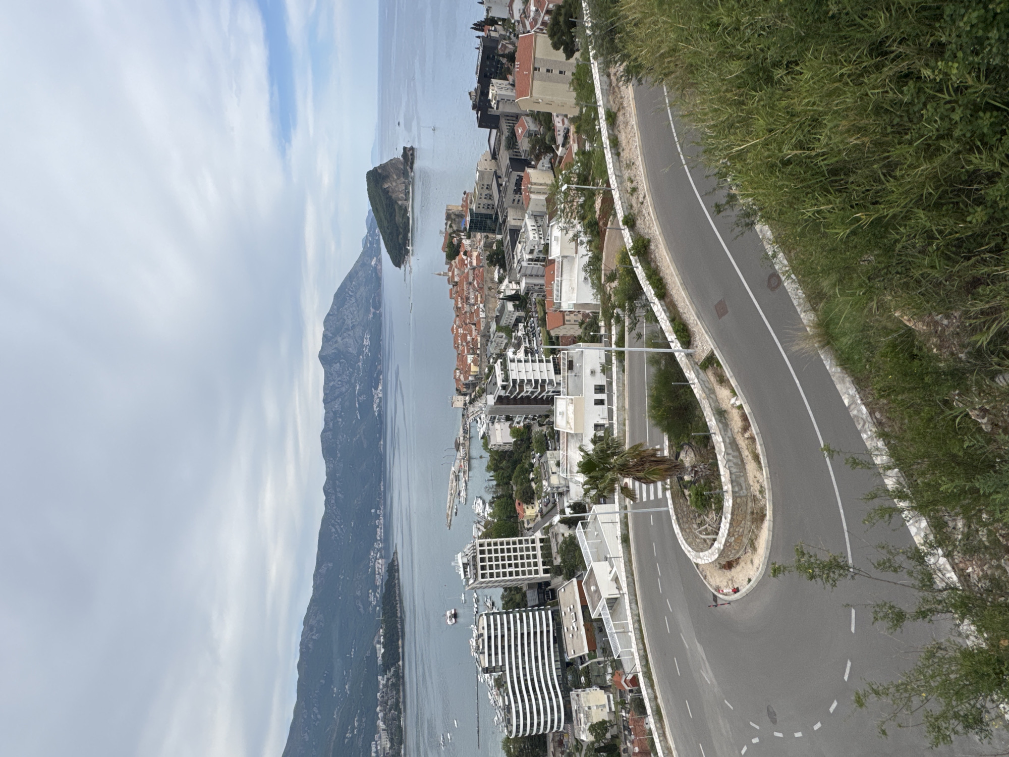 A high-angle, panoramic view captures a picturesque coastal town, identified as Budva, Montenegro, nestled between mountains and the sea. The foreground features a winding asphalt road with white markings, carving its way along a hillside. Dense green vegetation, including tall grasses, bushes, and a few trees (one palm tree is visible), lines the road and covers the surrounding slopes.
The middle ground is dominated by the sprawling town, stretching along the coastline. It comprises a mix of architectural styles, with many buildings featuring white or light-colored facades, some topped with traditional red-tiled roofs. Modern multi-story apartment buildings, including one with distinctive black and white striped patterns, are also visible. A calm body of water, likely a bay of the Adriatic Sea, lies adjacent to the town, dotted with numerous boats and yachts.
A prominent, verdant island, likely Sveti Nikola, rises from the sea in the middle distance, adding a striking natural feature to the landscape. Beyond the town and the water, a range of majestic, rugged mountains forms a dramatic backdrop. The sky above is partially cloudy, displaying shades of light blue with scattered white clouds, suggesting a pleasant daytime setting, likely late morning or early afternoon, with bright but diffused natural light. No specific human activity is discernable, nor is there any visible text. The overall scene exudes a peaceful and scenic atmosphere.