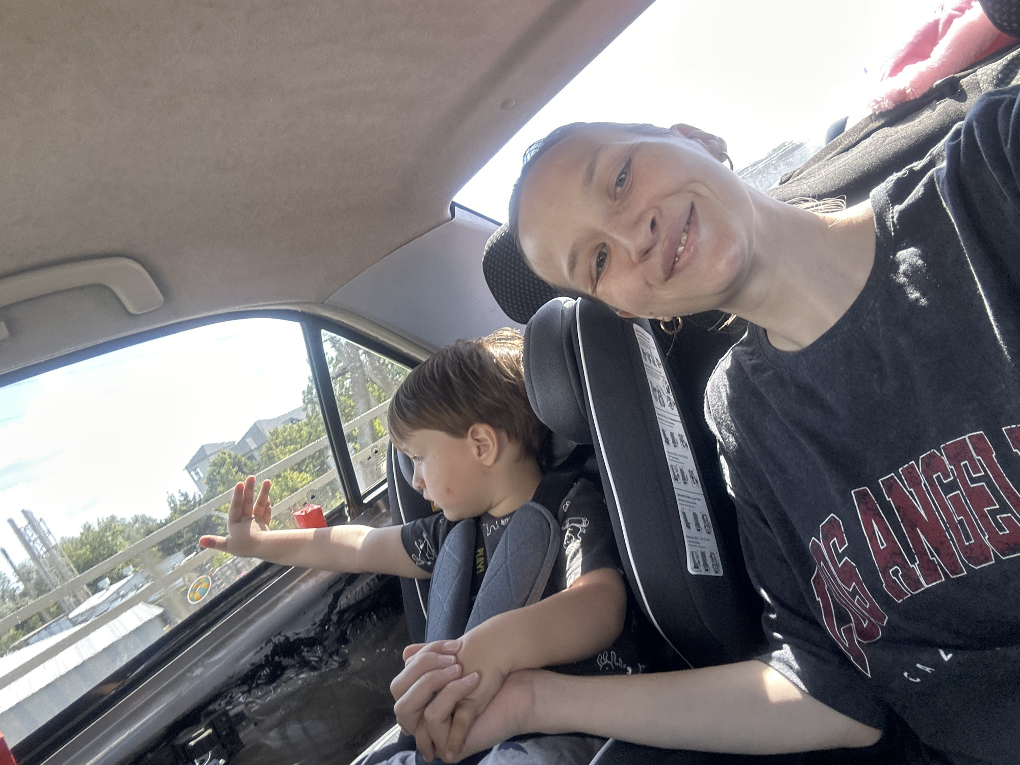 A woman and a young child are in the back seat of a car. The woman is in the passenger seat, taking a selfie. She is smiling and looking at the camera. The child is in a car seat next to her, looking out the window. The car is moving, and the scenery outside is blurred. The interior of the car is visible, including the seats, the ceiling, and the windows. The woman is wearing a dark t-shirt with "LA" printed on it. The child is wearing a dark t-shirt with a cartoon print. The scene is captured on a sunny day. The image is a candid shot, likely taken during a car ride. No information about the specific location in Peresyp, Russia is available from the image itself.
