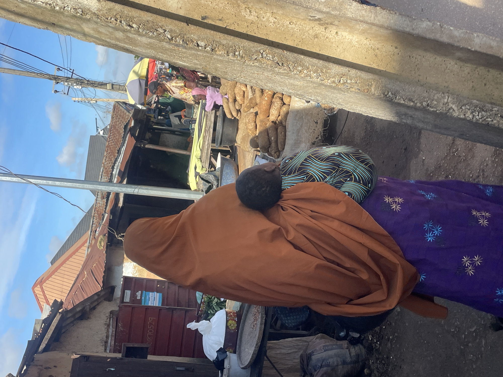 In a market scene in Jos, Nigeria, a woman wearing a burnt orange hijab and a purple skirt embroidered with blue and gold floral patterns carries a child on her back. The child is partially visible, wearing a patterned shirt with blue and green wavy lines. They are standing on a dirt path in front of market stalls. To the left, a stall displays various items, including a white bag on top of a circular object with colorful patterns and a woven tray with small brown objects. In the background, market structures with corrugated metal roofs are visible. Further back, other market vendors and shoppers are present, including a person in a light-colored patterned shirt and a woman in a pink headscarf and top with a child. Piles of yams are stacked on the ground in front of one stall. Overhead, power lines crisscross a bright blue sky with scattered white clouds. A rough, concrete structure runs along the right side of the frame, partially obscuring the market scene.