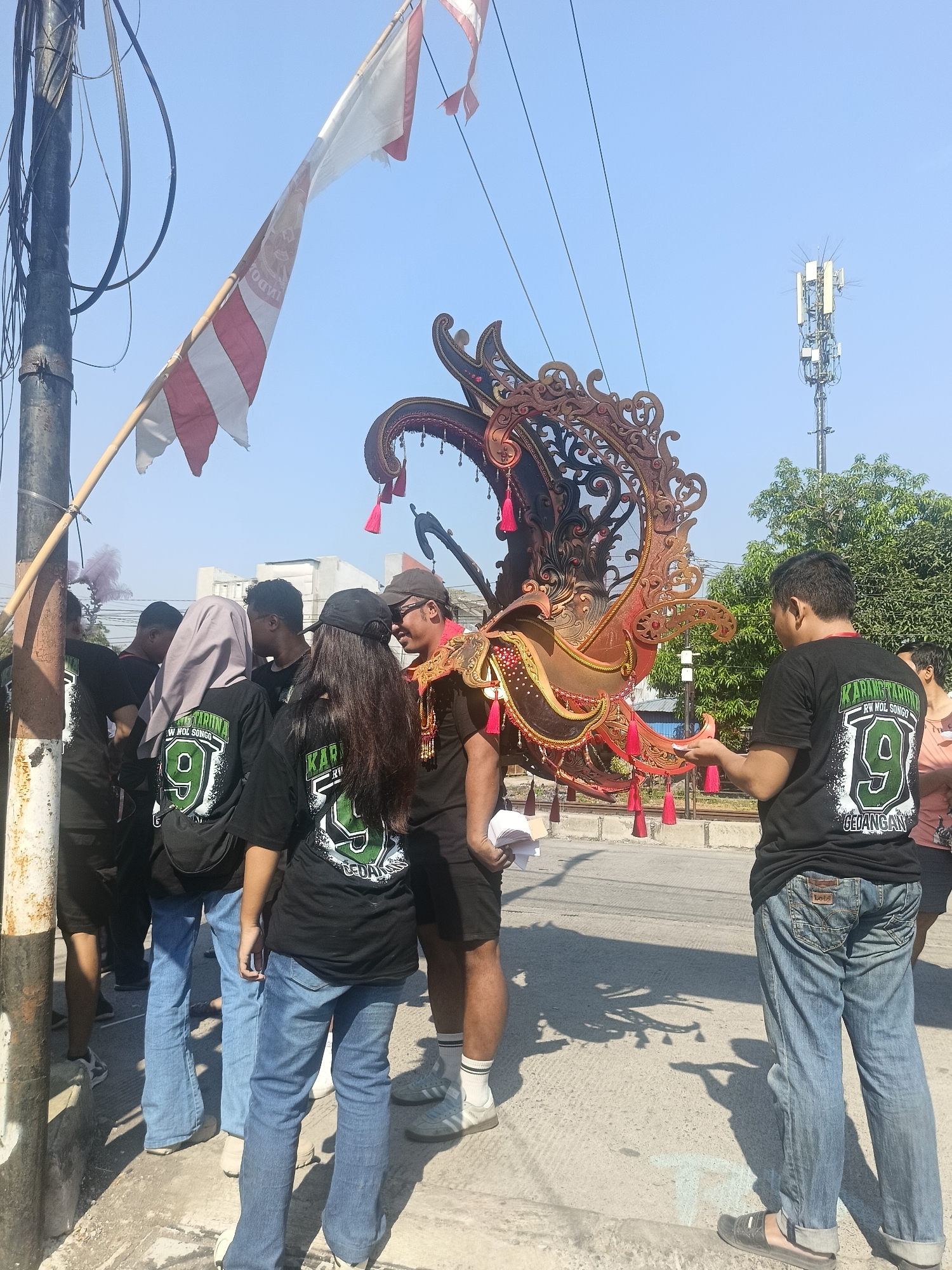 The image captures an outdoor scene on a bright, sunny day in Gedangan, Indonesia. In the foreground, a group of young adults are gathered near a street, facing a large, ornate decorative structure. The structure appears to be made of wood or metal, with intricate carvings and adorned with red tassels and gold accents. Several individuals are wearing matching black t-shirts with green lettering and a graphic that includes the number "9". One person in the foreground, a man, is wearing a t-shirt with the text "KARANGTARUNA RW NOL SONGO" and "GEDANGAN" visible. He is holding some papers in his hand. Another person, a woman, is also wearing a similar t-shirt and is looking towards the decorative structure.
To the left of the group, a utility pole with numerous wires is visible, and a red and white flag is attached to it, fluttering in the breeze. In the background, trees, buildings, and a cellular tower are present, indicating an urban or semi-urban environment. The ground is paved with concrete, and shadows from the people and the structure are cast on the surface. The overall atmosphere suggests a local event or celebration, possibly a parade or festival, taking place in the community of Gedangan.