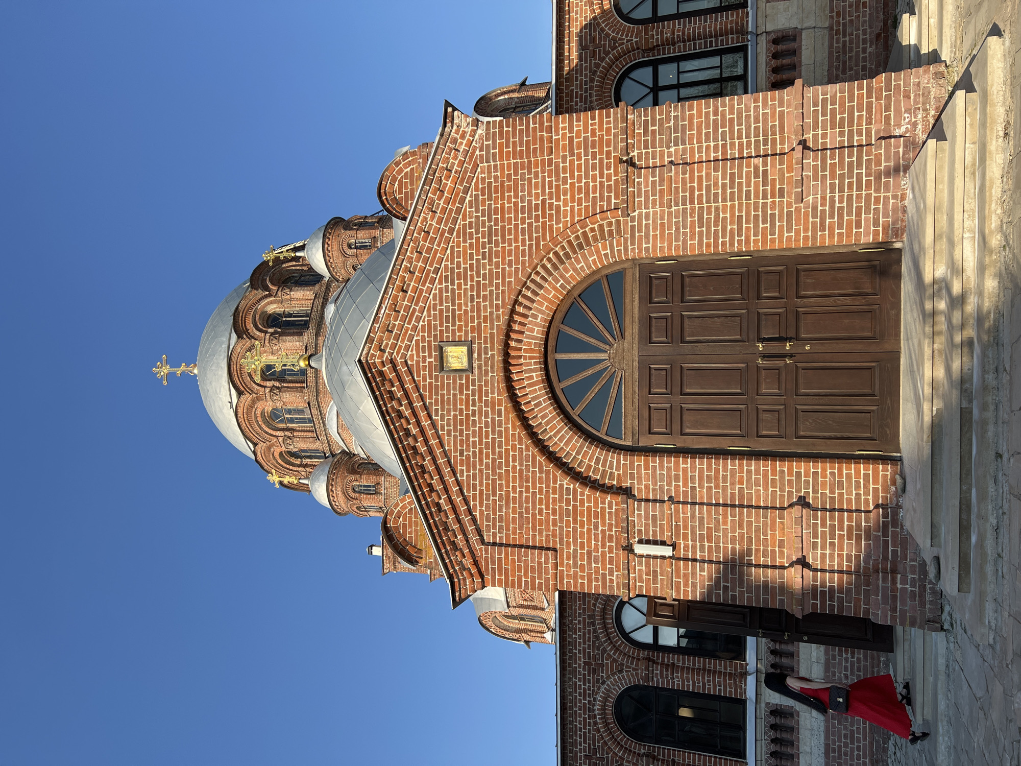 The image shows the red brick exterior of the Church of St. Constantine and Helena in Sviyazhsk, Russia. The building features arched windows and a large wooden door with a semicircular window above it. The church is topped with a silver dome and gold cross. A person in a red dress stands near the base of the building. The sky is clear blue.