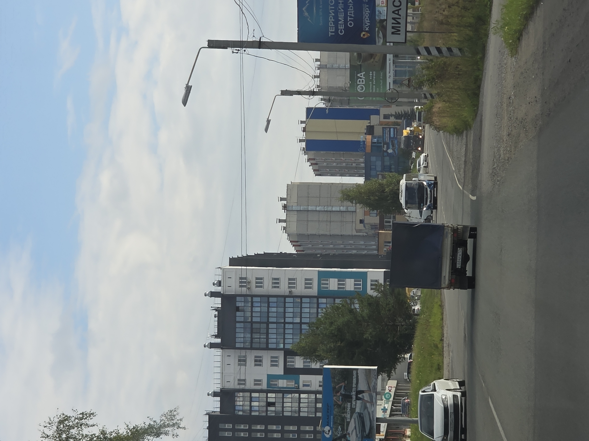 The image depicts a street scene in the city of Miass, Russia, under a cloudy sky. A white Volkswagen Polo is traveling on the right side of the road, while a blue-canopied truck is in the lane to its left. In the background, several multi-story apartment buildings are visible, with a bus and other vehicles on the road. There are also billboards and streetlights in the scene. The overall environment suggests an urban setting with ongoing traffic.