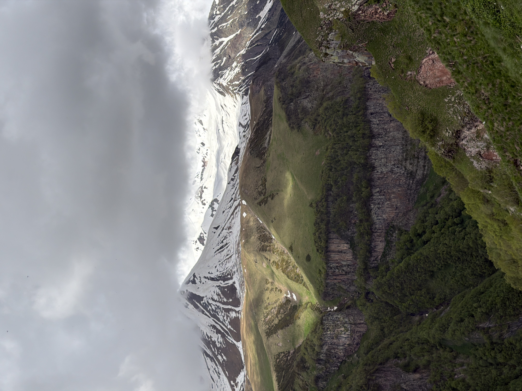 This is a panoramic, high-angle shot of a mountainous landscape in Gudauri, Georgia, likely taken in spring or early summer. The scene is dominated by dramatic geological formations and a varied terrain. The uppermost portion of the image features snow-capped mountain peaks partially obscured by thick, dramatic grey clouds. The midground showcases rolling green hills interspersed with patches of snow and rugged, rocky outcrops. Sunlight breaks through the clouds in places, illuminating sections of the slopes with a warm glow. In the foreground and lower sections, steep, verdant valleys are visible, densely covered with lush green trees and vegetation. The overall impression is one of wild, untamed natural beauty, with a stark contrast between the snow-covered peaks and the vibrant green of the lower elevations. There are no people or discernible man-made structures in the image, suggesting a remote, natural setting. The lighting is moody due to the cloud cover, with shafts of sunlight creating a dramatic interplay of light and shadow.