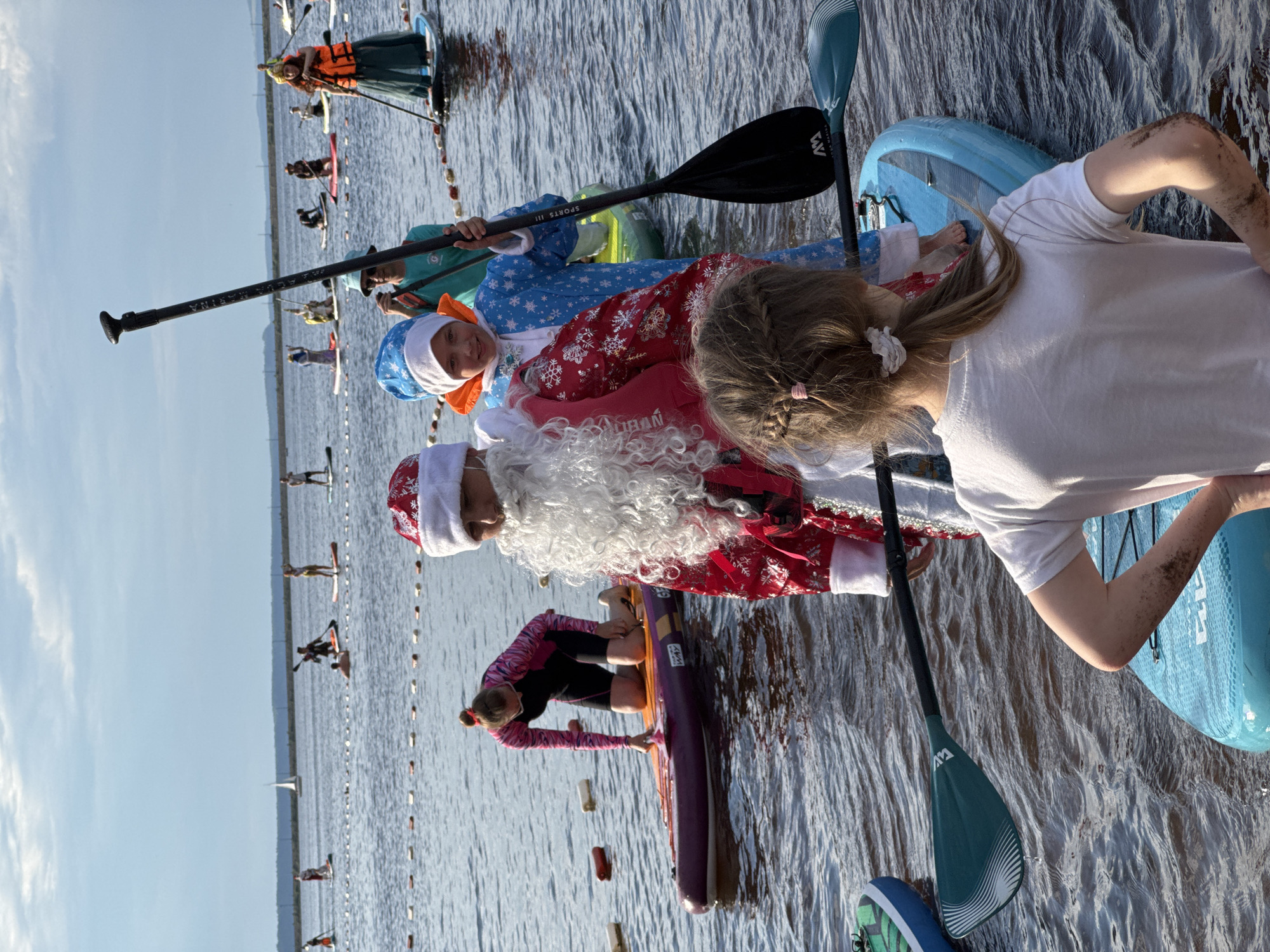 In Chaikovsky, Russia, a group of people are engaged in a paddleboarding event on a body of water under a cloudy sky. In the foreground, a person dressed as Santa Claus, complete with a red suit and white beard, stands on a paddleboard. To their left, a child in a blue and white snowflake-patterned costume, wearing a matching hat, also stands on a paddleboard and holds a paddle. Another child, seen from behind and wearing a white t-shirt, is on a blue paddleboard in the very front. In the background, numerous other individuals are paddleboarding, some in festive attire. A few sailboats are visible on the horizon, along with a distant shoreline. The water is calm, with a rope barrier marked by buoys indicating a designated area on the water.
