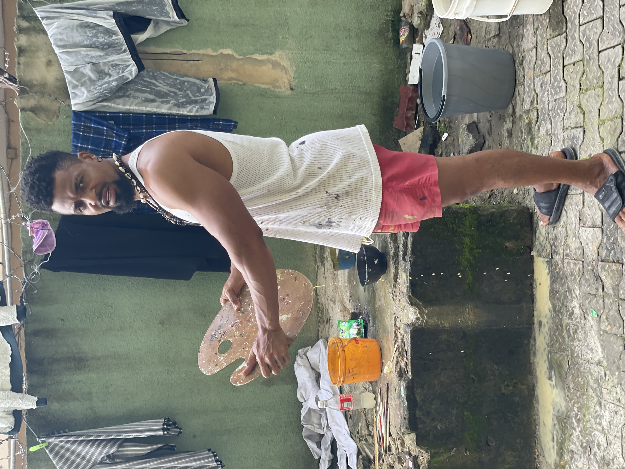 The image depicts a man standing in an outdoor setting, possibly an alleyway or a backyard in Ado, Nigeria. He is holding a wooden artist's palette, from which liquid is dripping onto a rough, uneven stone surface below. The man is wearing a white tank top splattered with paint, red shorts, and black slides. His left hand is on the palette, and his right hand is behind it.
The environment appears somewhat cluttered and utilitarian. To the left of the man, there is a raised concrete structure with various items on it, including a bottle of clear liquid, an orange bucket, and several paintbrushes. A black bowl sits on the ground near the orange bucket. In the background, a green-colored wall serves as a backdrop, with laundry hanging on a line above it. Various items of clothing, including striped shirts and what appear to be trousers or shorts, are visible on the line. The ground is paved with interlocking blocks and shows evidence of wetness and pooling water, with a yellowish liquid seeping from the stone structure. The overall impression is one of a working artist's informal outdoor studio space.