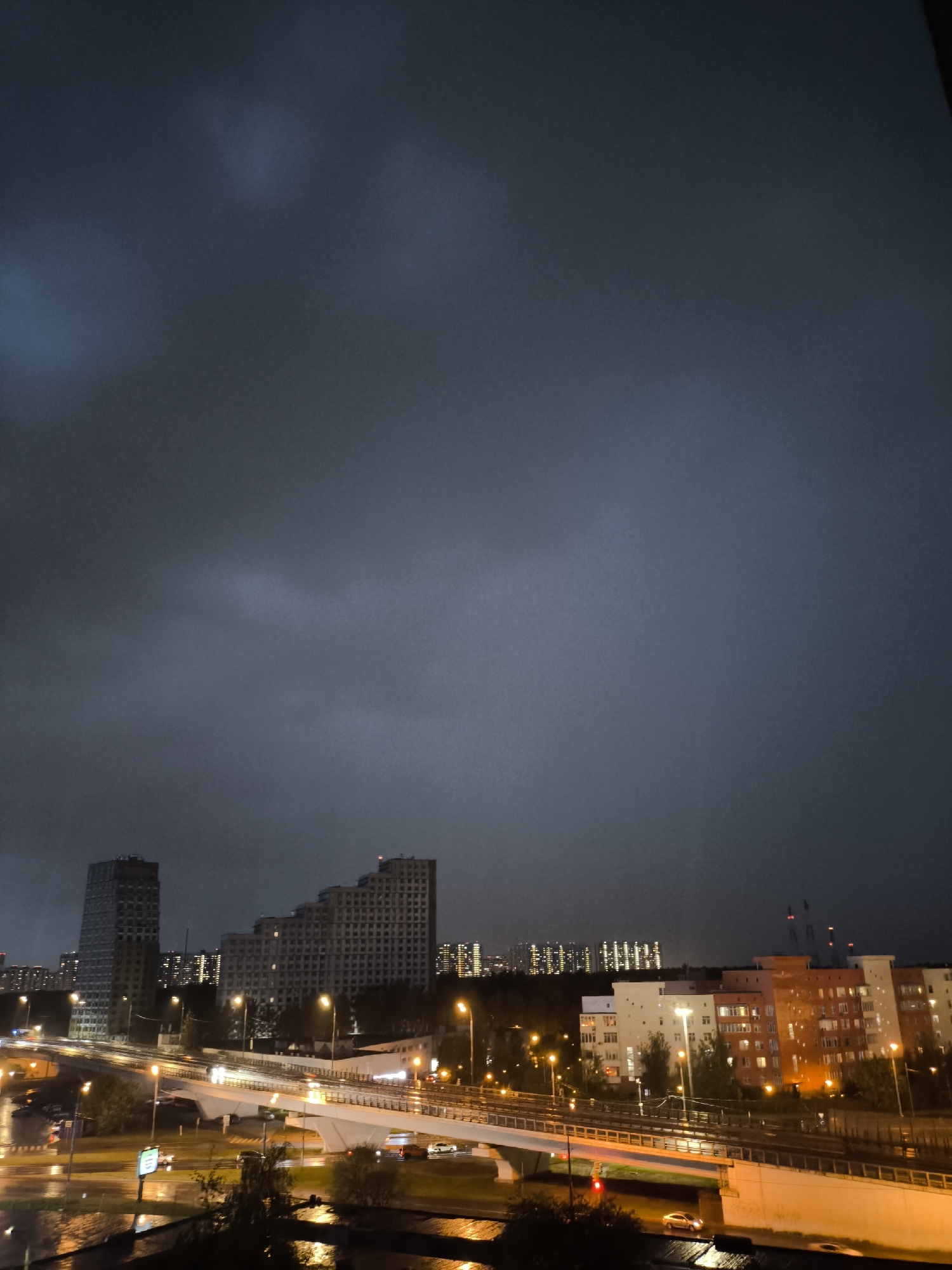 The image depicts a cityscape at night, likely after a rainstorm, as evidenced by the wet surfaces reflecting streetlights and car headlights. Dominating the skyline are several tall buildings, including a modern tower and a stepped apartment complex. A highway overpass is visible in the foreground, carrying traffic and illuminated by streetlights. In the distance, other residential buildings are visible. The sky is overcast with dark, heavy clouds. The location is Unknown, Unknown.