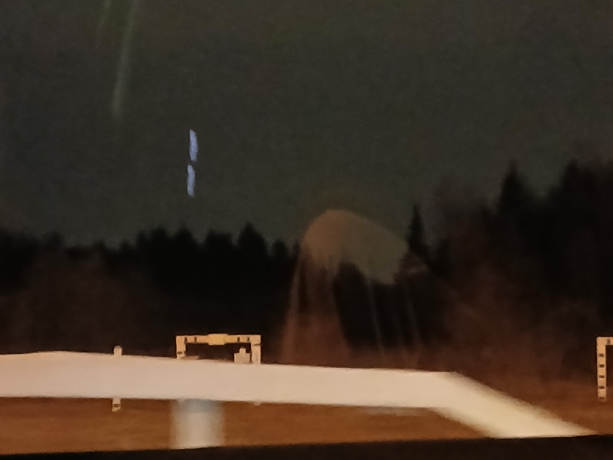 An outdoor nocturnal scene, likely captured from a vehicle, depicts an expansive, flat surface covered in white material, possibly snow or ice, under low artificial lighting. In the mid-ground, a light-colored hurdle-like structure with a horizontal bar and two vertical supports is visible on the left, while a segmented vertical post stands on the far right. A dense, dark treeline forms the background. The sky is dark with a faint, elongated, bluish-white vertical light streak on the upper left and a large, diffuse, yellowish-white glow emanating from the mid-right, possibly a reflection or an external light source. No individuals are present. Location: Unknown, Unknown.