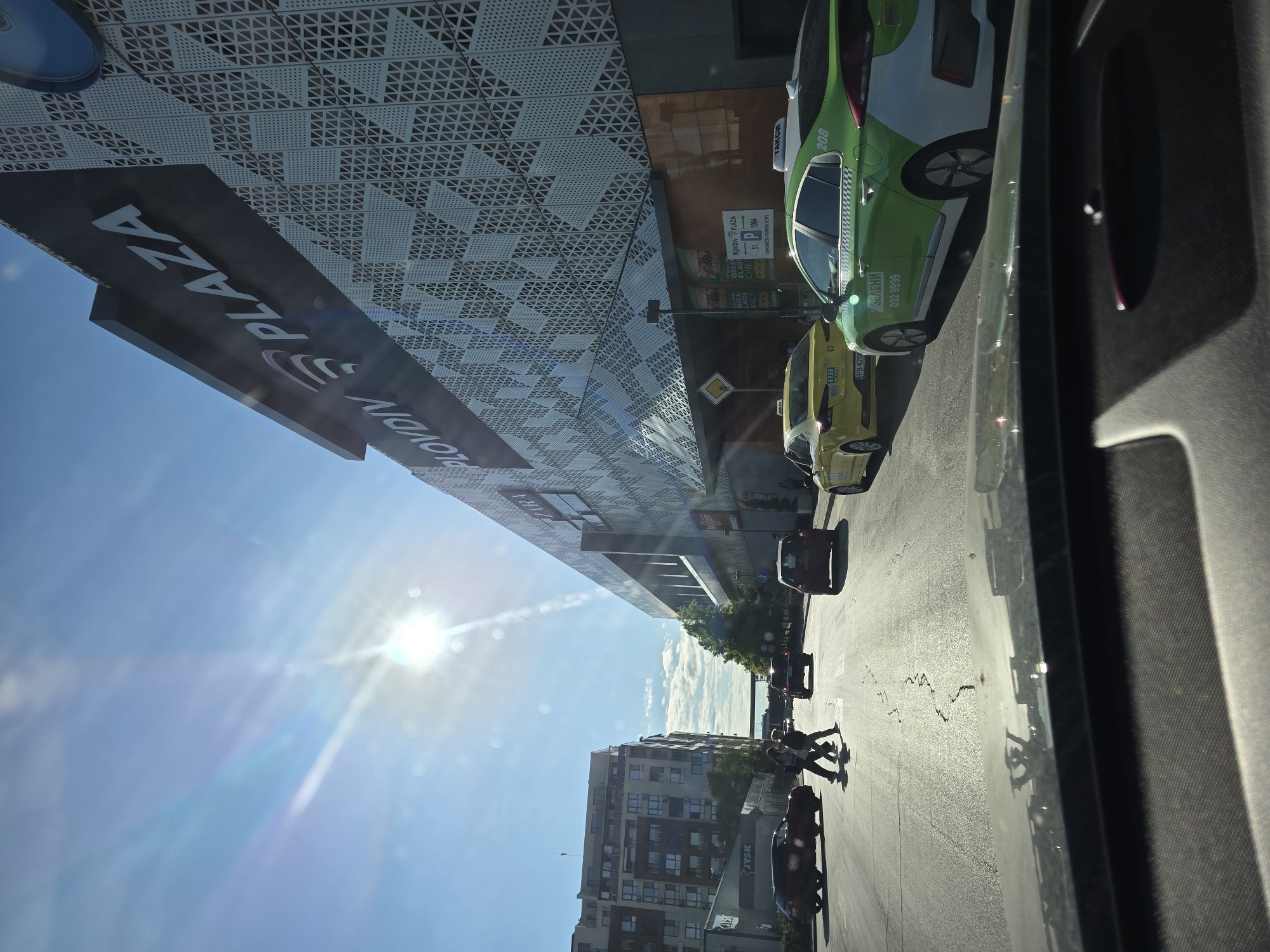 The image depicts an urban street scene in Plovdiv, Bulgaria, captured from inside a vehicle on a bright, sunny day. The perspective is angled towards a modern, multi-story building on the left, which features a distinctive facade composed of geometric, perforated panels.
A large black sign on this building prominently displays "PLOVIN PLAZA" in white capital letters. Below it, a smaller green sign includes the text "GREEN FLAGS ONL", "511004", and Cyrillic text that translates to "PARKOMESTA I PARKING SLOTOVE" (Parking spaces and parking slots).
On the street below, several vehicles are present. A green and white taxi, with the Cyrillic word "ТАКСИ" (TAXI), the phone number "032 9999", and the number "208" on its side, is parked near the building. Behind it, a yellow taxi is visible, marked with "6155" and a Bulgarian license plate "PB 420P". Further down the street, a dark red car is also visible. A yellow diamond-shaped sign, indicating a "priority road", is on the street.
A few pedestrians are seen on the sidewalk and crossing the street, their long shadows stretching across the asphalt, suggesting it is mid-day to early afternoon. The sky is clear and blue, with the bright sun creating a noticeable lens flare in the upper left corner of the image. In the background, other buildings line the street, including one with a "JYSK" sign visible. The scene portrays a bustling city environment under a clear sky.