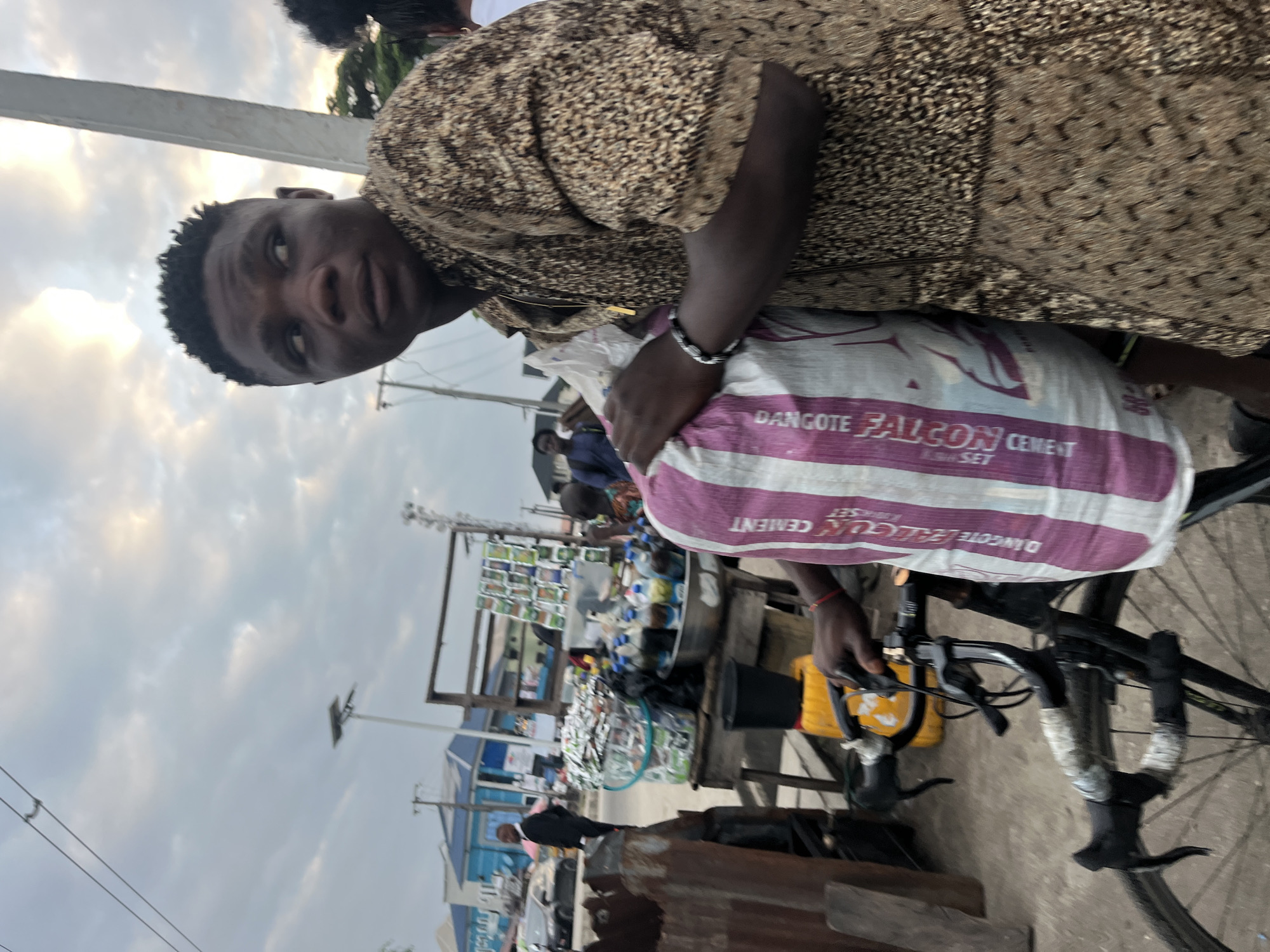 A young Black male, appearing to be in his late teens or early twenties, stands in what appears to be a bustling outdoor market scene in Ado, Nigeria. He is looking to his right, away from the camera, with his head turned. He wears a tan and brown patterned shirt and has short, dark hair. He is holding a large, striped bag with text in what looks like purple and white. The bag has the word "CEMENT" clearly visible on it, along with the brand "Dangote". The man's left arm is resting on the handlebars of a black bicycle, which is positioned in front of him. The bicycle appears to be a road bike, with its handlebars wrapped in white tape.
Behind the man and the bicycle, a market stall is visible. It is laden with various goods, including bottles of liquid, what appear to be small packages or sachets, and a large clear plastic container filled with more items. A man in a dark suit can be seen walking in the background, further down a street lined with buildings, one of which is painted blue. The sky above is cloudy with patches of light, suggesting it may be late afternoon or early evening. The overall environment appears to be an urban street market.