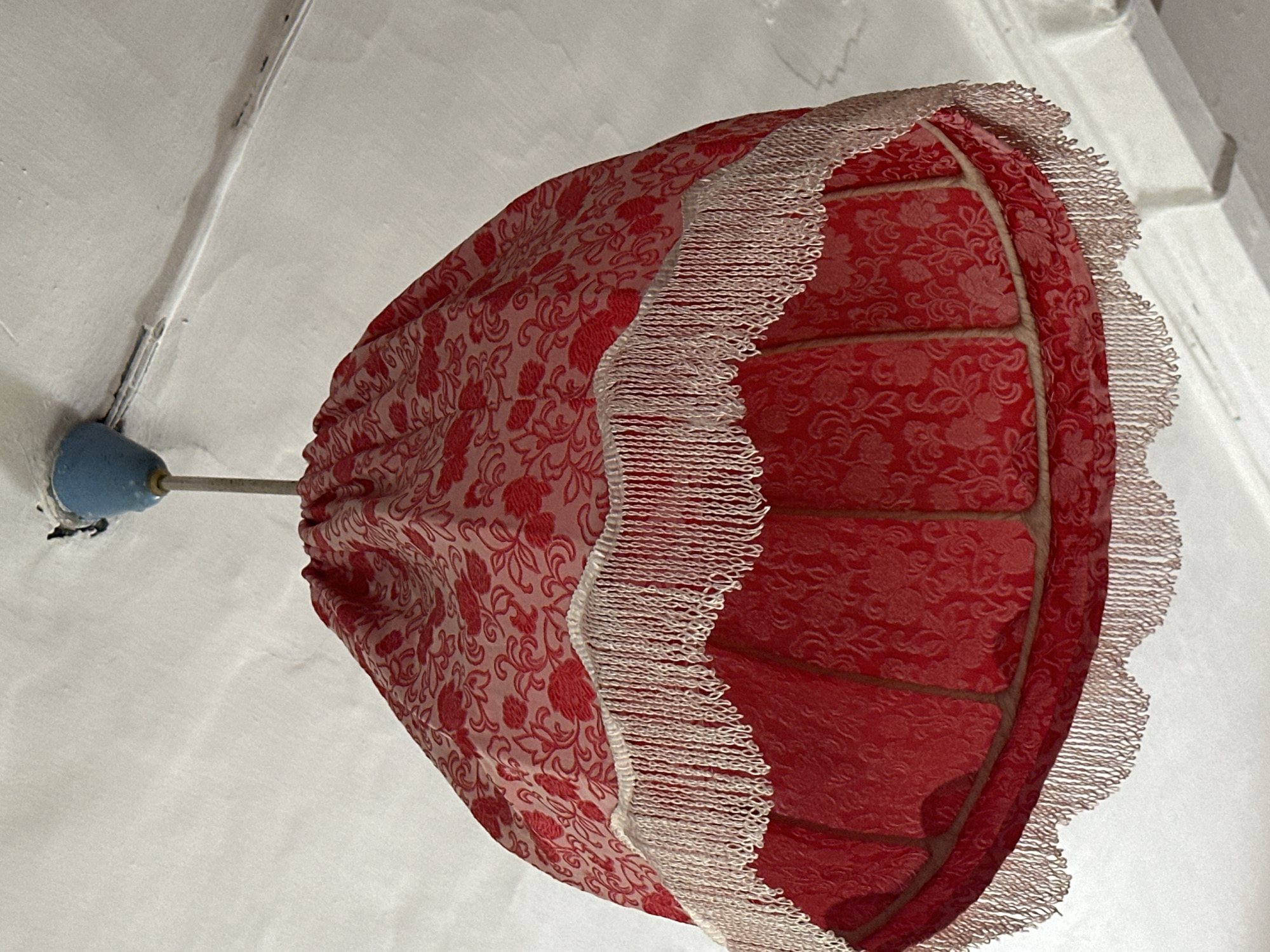 A close-up, upward shot shows a red fabric lampshade with a floral pattern, adorned with white fringe along its lower edge. The lampshade is suspended from the ceiling by a thin metal rod, attached to a blue plastic fixture. The ceiling itself is white and appears textured. The scene is indoors, likely in a residential setting. The location is not discernible from the image itself.