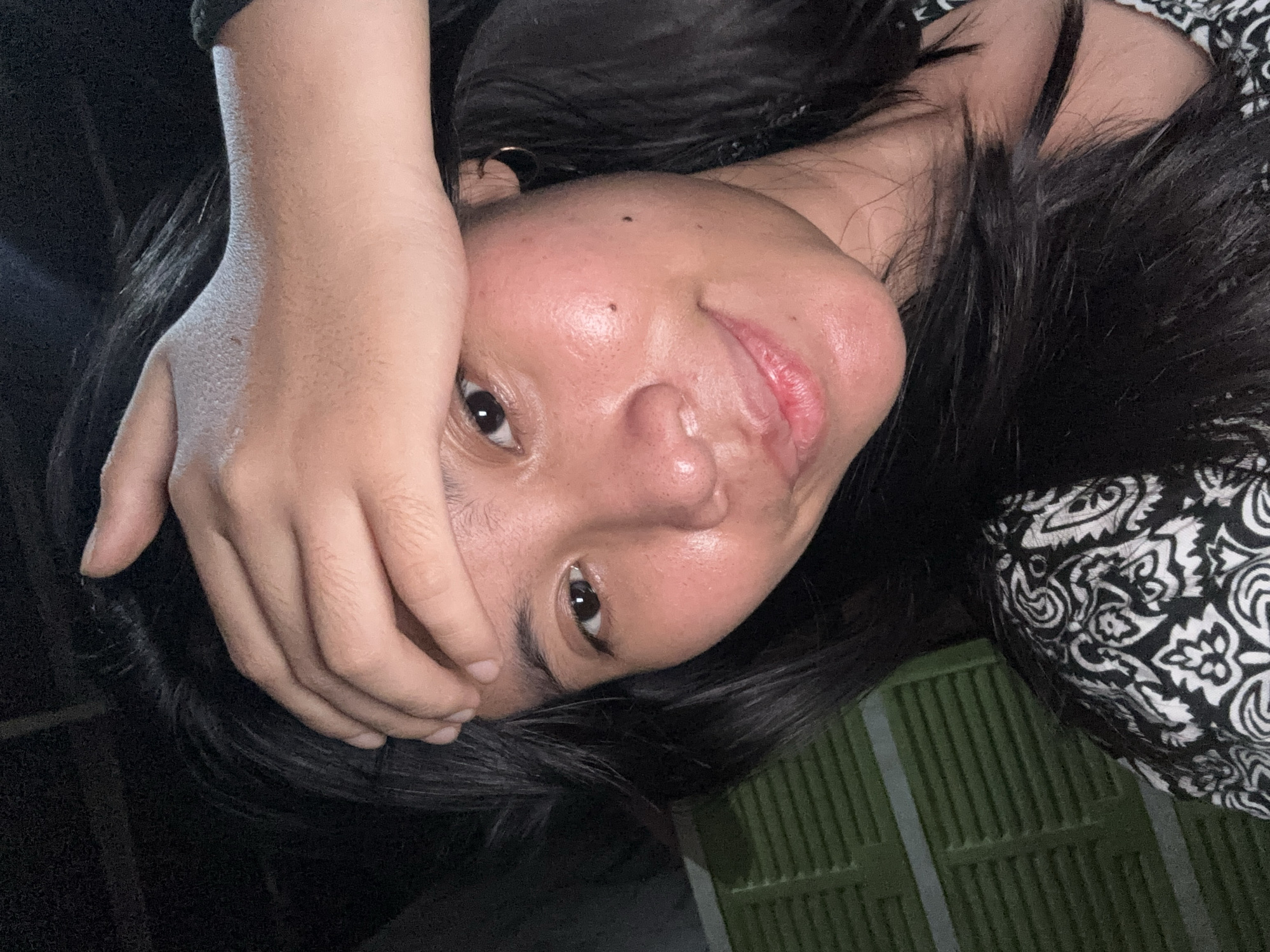 This media file is a close-up, rotated selfie of a person. The person has dark hair and fair skin. Their right hand is placed over their forehead, obscuring part of their face. One eye is visible, looking towards the camera. Their lips are slightly parted, and their mouth appears moist. There is a small dark spot on their forehead, possibly a mole. They are wearing a black garment and a garment with a black and white patterned design. The background includes a dark area and a green object with a ribbed texture, possibly a plastic crate or a textured wall. The lighting suggests it might be indoors or during the evening. The context provided indicates the location is East Jakarta, Indonesia.