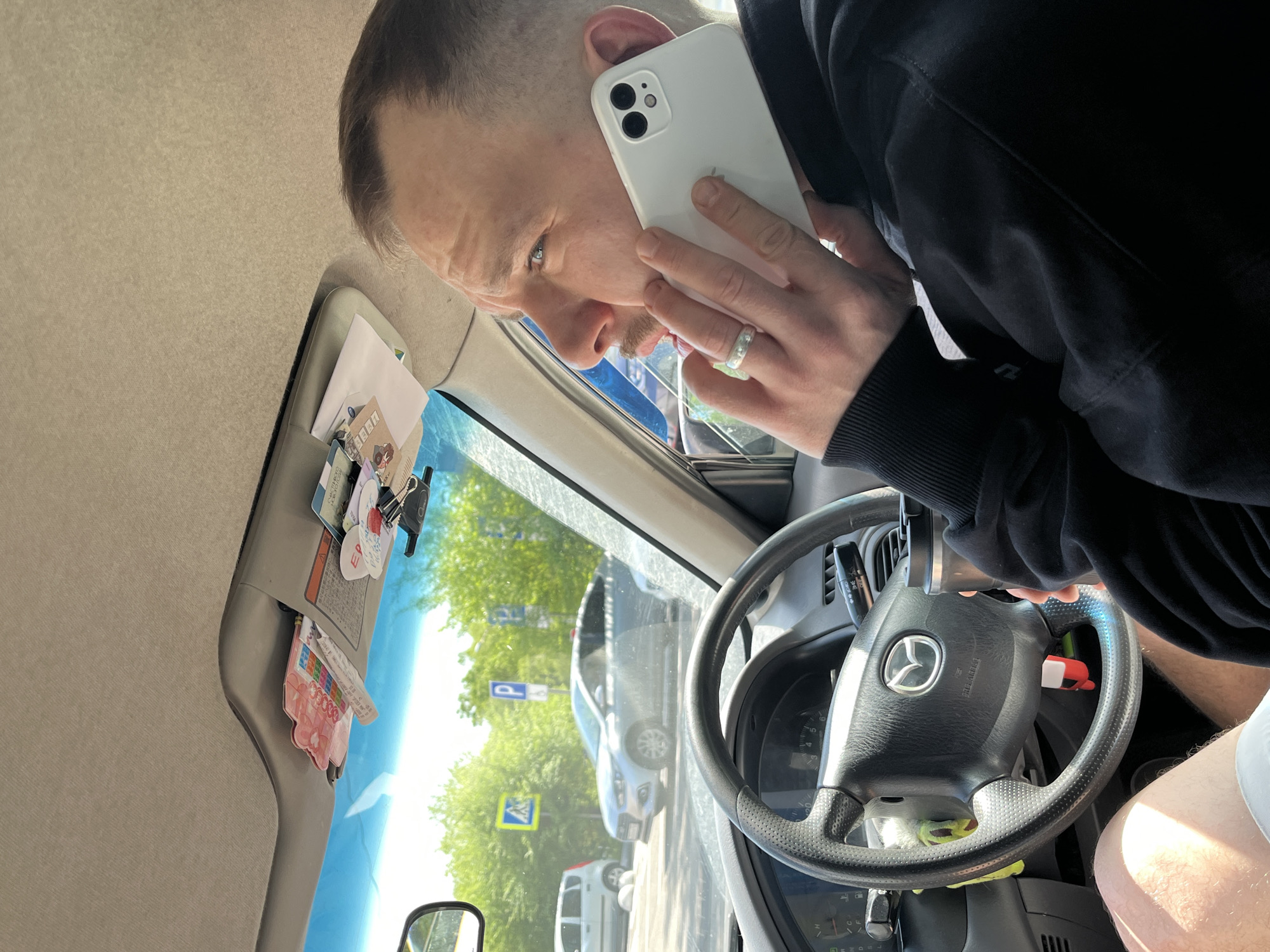 A man with short hair is in the driver's seat of a car, holding a white smartphone to his ear. He is wearing a black hooded sweatshirt. The steering wheel has a Mazda logo. The car's sun visor is down and has various cards and papers tucked into it. Outside the car, there is a parking sign and trees, suggesting a parking area. Based on the parking sign and the architecture visible through the windshield, the location is identified as the city of Krasnoyarsk, Russia.