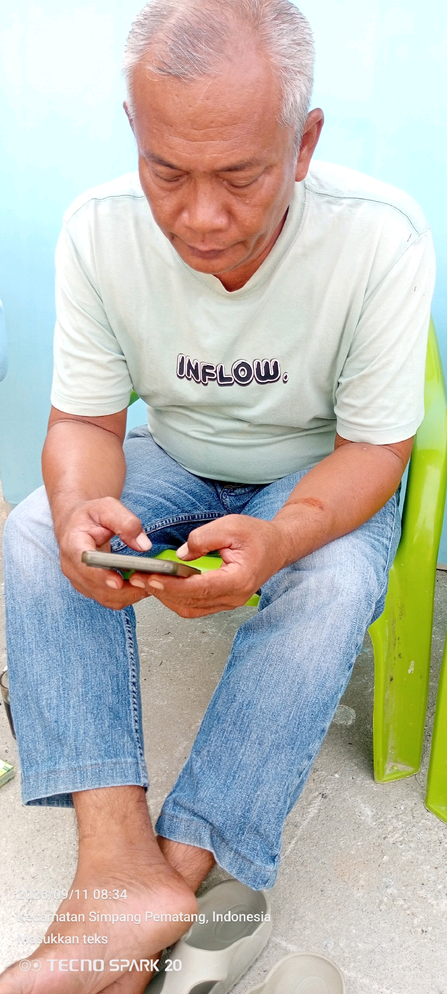 The image shows a man sitting on a bright green plastic chair outdoors, looking down at a smartphone he is holding in his hands. He is wearing a light-colored t-shirt with the word "INFLOW." printed on it and blue jeans. His feet are bare. The background includes a light blue wall and a concrete floor. A timestamp and location overlay at the bottom of the image reads "2026/09/11 08:34 Kecamatan Simpang Pematang, Indonesia".