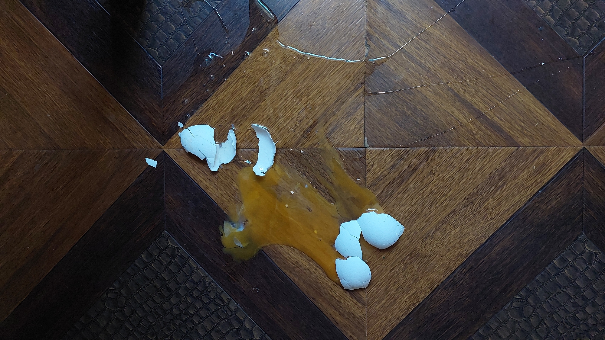 A cracked egg is spilled on a hardwood floor. The egg yolk and white are spread across the surface. Fragments of the eggshell are scattered around the spill. The floor is made of dark and light brown wooden planks arranged in a diamond pattern. There are textured dark brown sections at the edges of the frame. The scene is indoors.