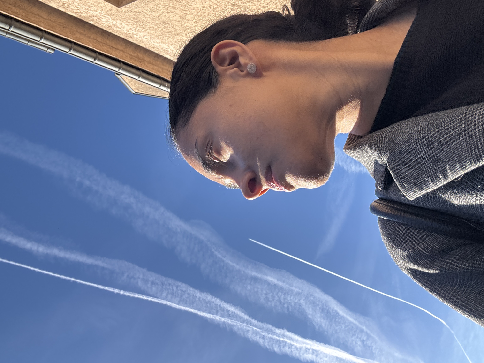 A young woman, viewed from the side, is looking upwards at a bright blue sky filled with contrails from airplanes. She has dark hair pulled back into a ponytail and is wearing a grey plaid jacket over a dark top. A diamond stud earring is visible in her right ear. The sun casts a warm glow on her face, highlighting her features. To the right of the woman, the upper portion of a light brown building with a grey gutter is visible, indicating an urban or suburban setting. The image appears to have been taken in Pétange, Luxembourg, based on the context provided.