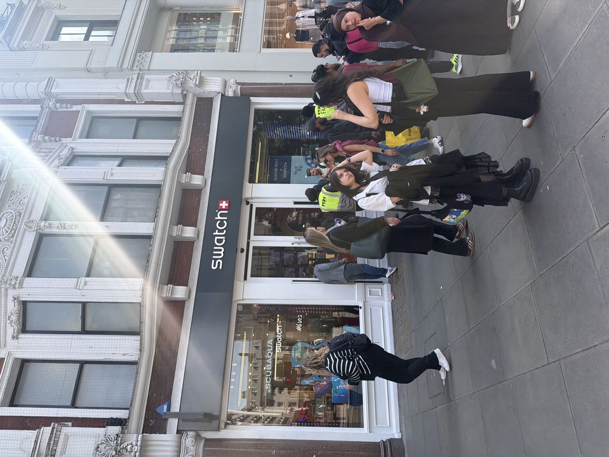 A street-level exterior shot shows a Swatch store with a crowd of people gathered outside. The store has a black and white Swatch sign and window displays. A sign on the glass says "SCUBAQUA" and "#huna". The people are dressed in a variety of casual clothing and are standing and talking. The architecture of the building features white ornate details and a mix of brown and white brick. The sidewalk is paved with gray stone. The image suggests the store is on Oxford Street in London, United Kingdom, given the presence of an "AUDEMARS PIGUET" sign and the architectural style characteristic of the area.