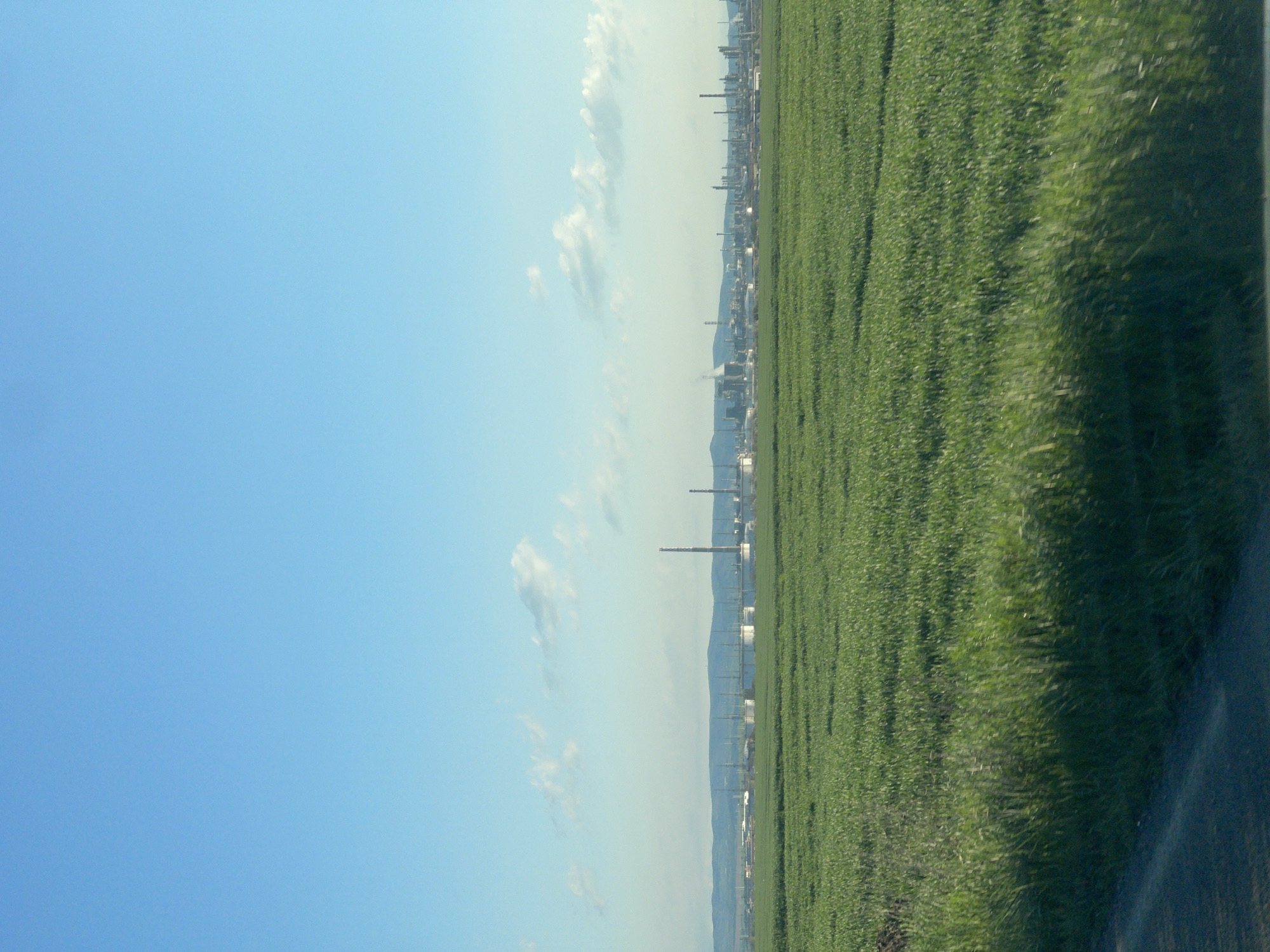 The image shows a field of green crops, likely grass, adjacent to a road. In the distance, a city skyline is visible, interspersed with industrial structures, including smokestacks. The sky is light blue with scattered clouds. The scene is likely located near Bratovo, Bulgaria. There are no people visible in the image.
