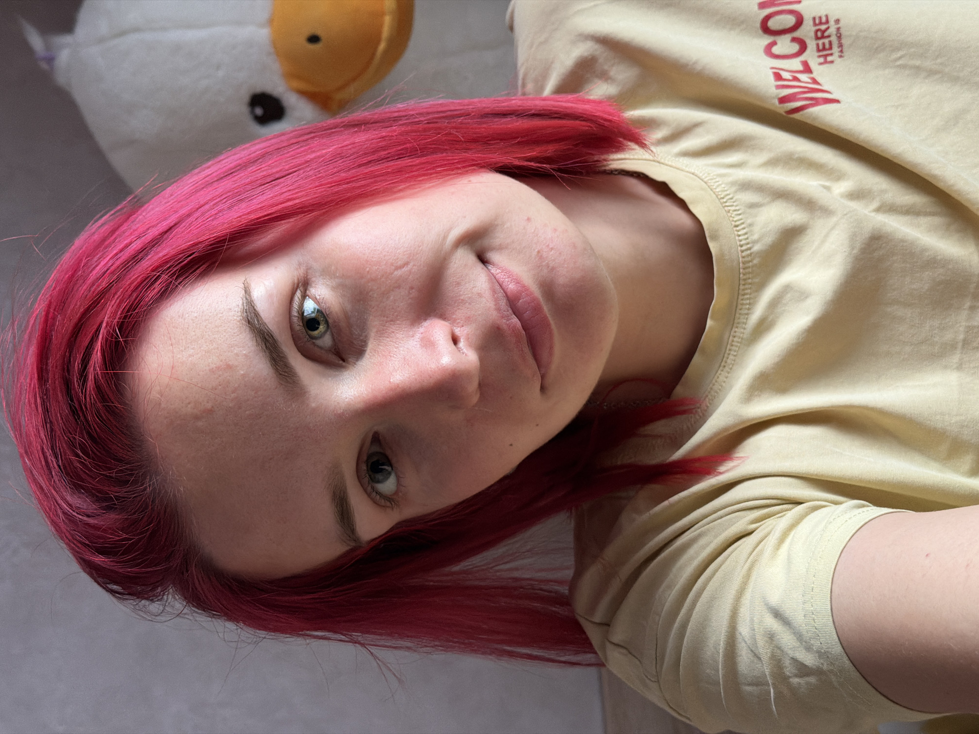 A young woman with vibrant pink hair and green eyes looks directly at the camera, smiling slightly. She is wearing a light yellow t-shirt with red lettering that says "WELCOME HERE FASHION IS". Behind her, out of focus, is a large, plush white duck toy with a yellow beak. The background appears to be a plain wall. The image seems to be a selfie taken indoors, possibly in a bedroom. There is no indication of the location being in Chaikovsky, Russia.