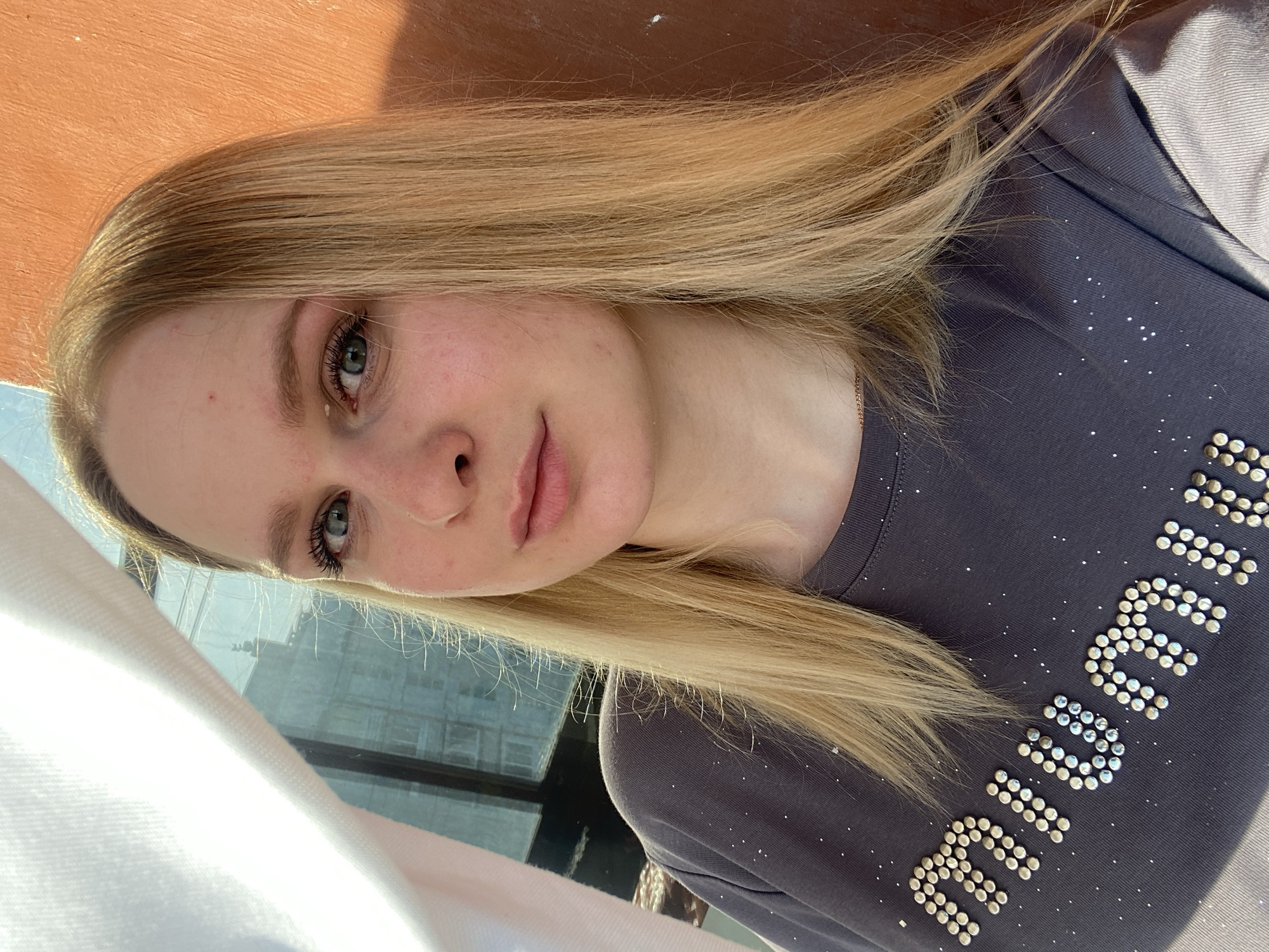 A fair-skinned young woman with blonde hair is pictured indoors. She is wearing a gray t-shirt with a pattern or design on it. The background includes a building visible through a window.