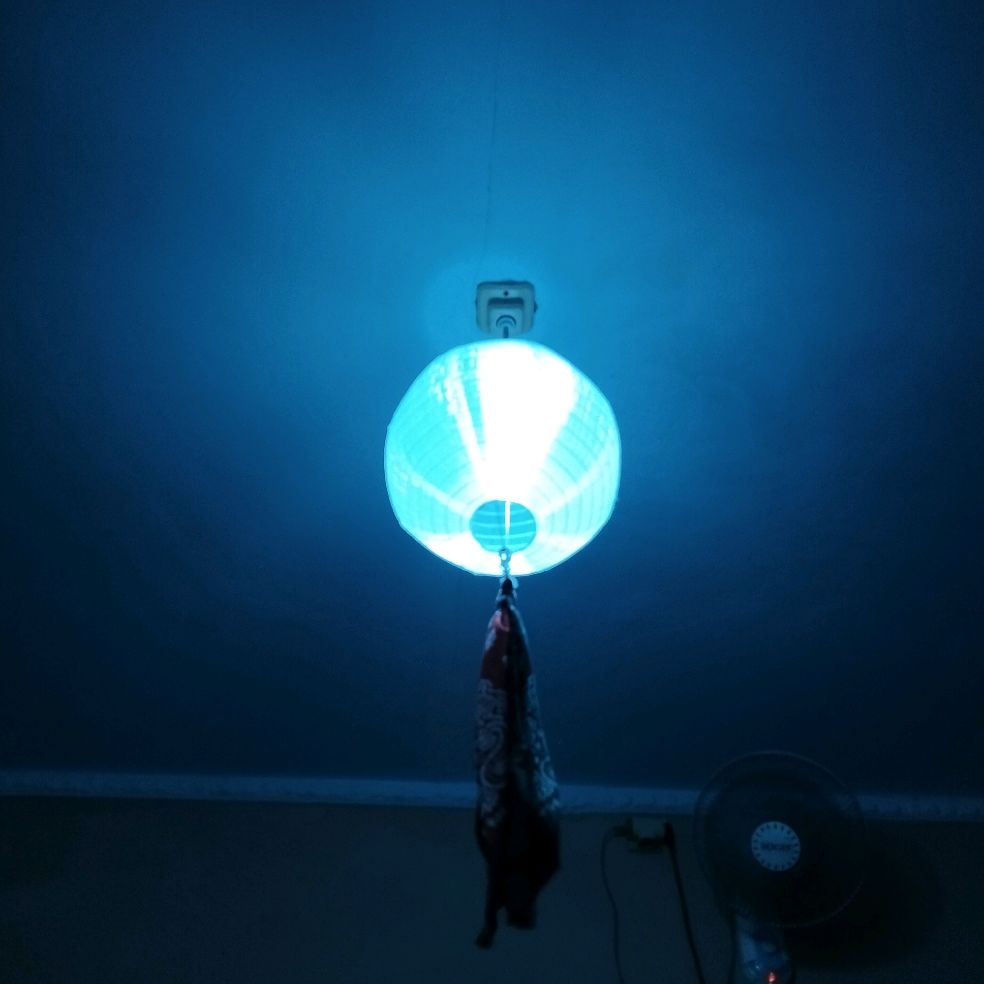 A close-up, low-angle shot captures a vibrant, aqua-colored paper lantern hanging from a ceiling, illuminated from within by a bright white light. The lantern's ribbed texture is visible, and a portion of a decorative tassel hangs from its bottom. Above the lantern, a white electrical fixture is attached to the ceiling. The surrounding ceiling is a deep, saturated blue, gradually fading to a darker shade towards the top and sides of the frame, suggesting ambient lighting. In the lower right corner, a dark, circular fan with a central logo is partially visible, and in the lower left, a light-colored electrical outlet with a black cord is present. The overall scene is dimly lit, with the bright lantern being the primary light source. The image does not provide any specific information about the location within Bandung City, Indonesia.