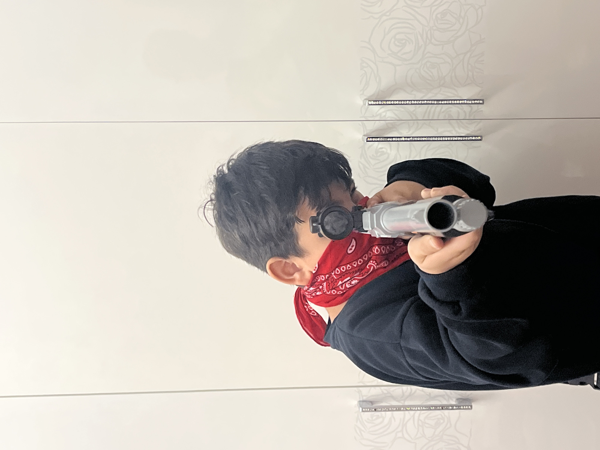 A child is aiming a toy gun in an indoor setting. The child is wearing a black sweatshirt and a red bandana around their neck and face. They are holding the toy gun with both hands, focused on aiming. The background consists of a white wardrobe with a subtle rose pattern and two decorative handles. There are no indications of the specific location within the city of Kostroma, Russia.