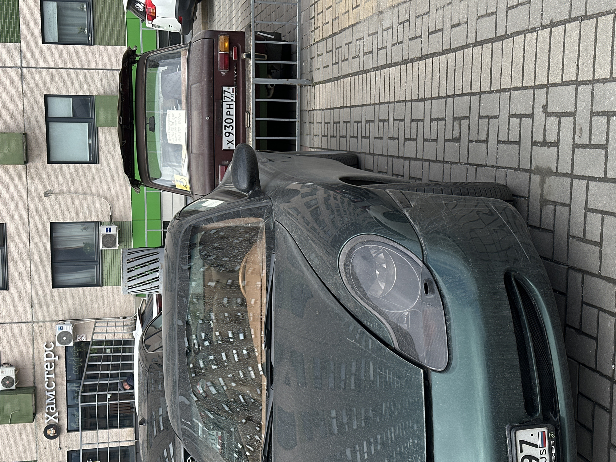 The image depicts a dusty green Alfa Romeo 8C Competizione parked near a brick sidewalk, with a rusty brown vintage Lada Riva car parked behind it. The Lada has a Russian license plate that reads "X930 PH 77" and a sign on the rear window. The Alfa Romeo also has a Russian license plate with partially visible characters. The photo was taken in Khimki, Russia as there is sign in Russian, "Хамстерс" (Hamsters) in front of the building in the background. The architectural style of the building indicates a typical urban setting in Russia. The image suggests the contrast between classic and modern car designs.