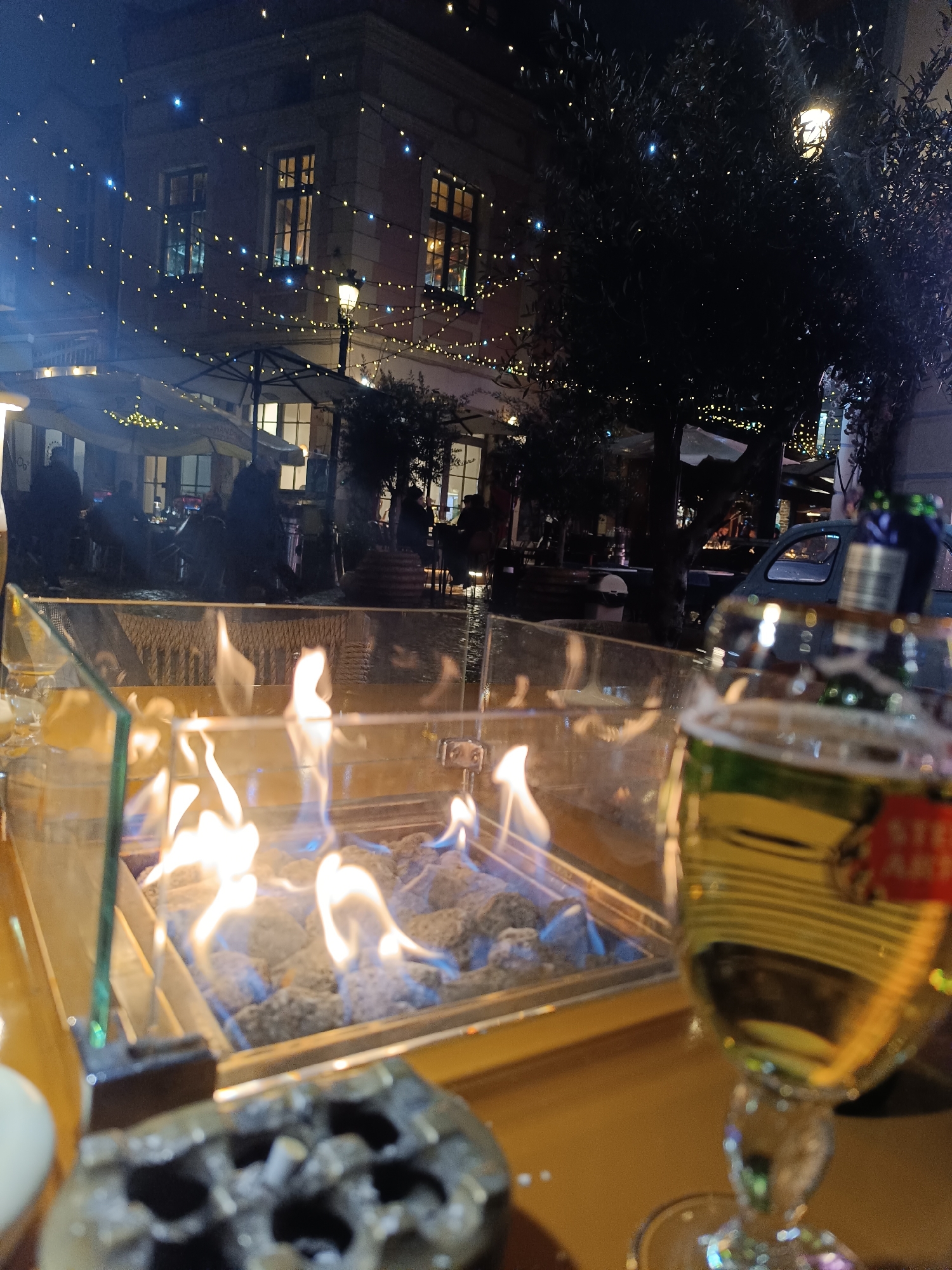 The image depicts a nighttime scene on a public square or pedestrian street in Plovdiv, Bulgaria. In the immediate foreground, a light-colored table features a rectangular, glass-enclosed fire pit with active blue and orange flames rising from grey decorative stones. To the right, a stemmed glass containing a yellow liquid, identifiable by a red and white Stella Artois label, is partially visible. A dark, metallic ashtray with ash and cigarette butts rests on the table. A white ceramic cup is partially seen on the far left.
Beyond the table, the midground reveals an outdoor seating area of a cafe or restaurant. Several indistinct human figures are seated at tables beneath large, light-colored awnings or umbrellas. Numerous yellow and blue string lights are strung horizontally across the scene, extending between buildings and dark-leaved trees. On the far right, a vintage light-blue car is parked.
The background consists of a multi-story building with illuminated windows, suggesting internal activity. A street lamp provides additional ambient light. The overall environment is an active, urban night setting characterized by social gathering and decorative illumination.