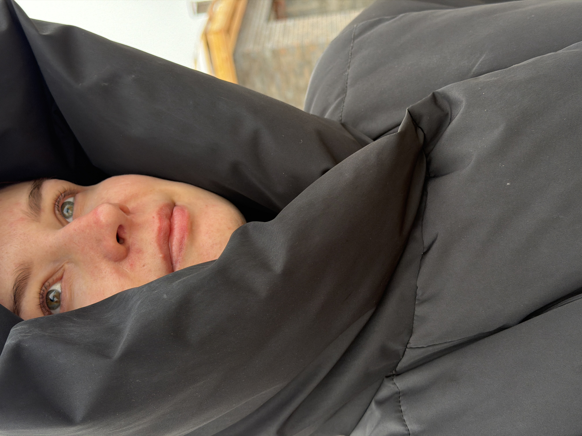 A person with light skin and green eyes is mostly obscured by a black hooded jacket. The left side of the person's face, including an eye, nose, and mouth, is visible. The hood of the jacket is pulled closely around the face. The location is suggested to be Chaikovsky, Russia. The image is shot from a low angle. The background is blurred, with indistinct shapes suggesting a building or outdoor structure.