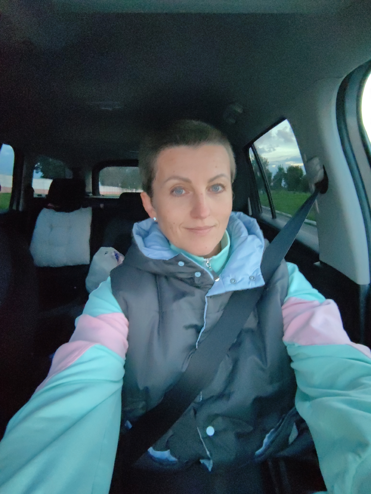 A selfie taken from the driver's seat of a car shows a woman with short, dark hair and blue eyes. She is wearing a puffy vest over a teal turtleneck and a jacket with teal and pink sleeves. A black seatbelt is fastened across her chest. The interior of the car is visible, with car seats and bags in the back. Through the windows, a landscape of green trees and a cloudy sky can be seen. The image was taken in the city of Myadzyel, Belarus.