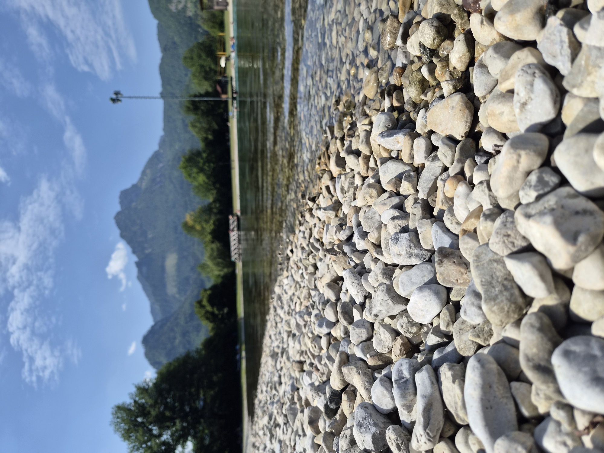 The image is a low-angle shot of a rocky shore on a sunny day. In the foreground, smooth, light-colored pebbles dominate the scene, with some wet stones near the water's edge. The water is clear and shallow, revealing more stones beneath the surface. In the midground, a body of water stretches out, with a wooden walkway or pier extending into it from the left. Several people are visible in the water and on the shore in the distance, enjoying a recreational activity. The background features a line of green trees and a prominent mountain range under a blue sky with scattered white clouds. A tall, metal light pole is visible on the right side of the image. The overall setting suggests a lake or a similar body of water near a mountainous region, likely a public recreational area. This scene is consistent with the landscape of Landl, Austria, a town known for its proximity to the Austrian Alps and its natural beauty.