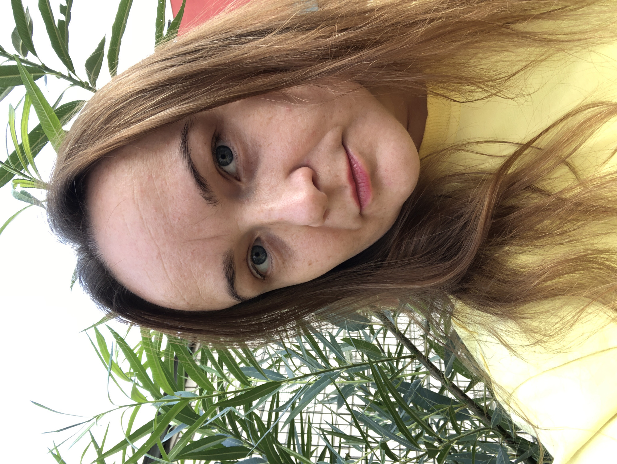 In the city of Bor, Russia, a medium-shot, outdoor, daytime photograph captures a woman with long, light brown hair, wearing a yellow top. She is positioned slightly off-center, facing the camera. Her facial features include blue eyes and light skin with freckles. Behind her, lush green foliage is visible, interspersed with some fencing, suggesting an outdoor environment possibly in a backyard or garden. The lighting is bright, indicating natural daylight.