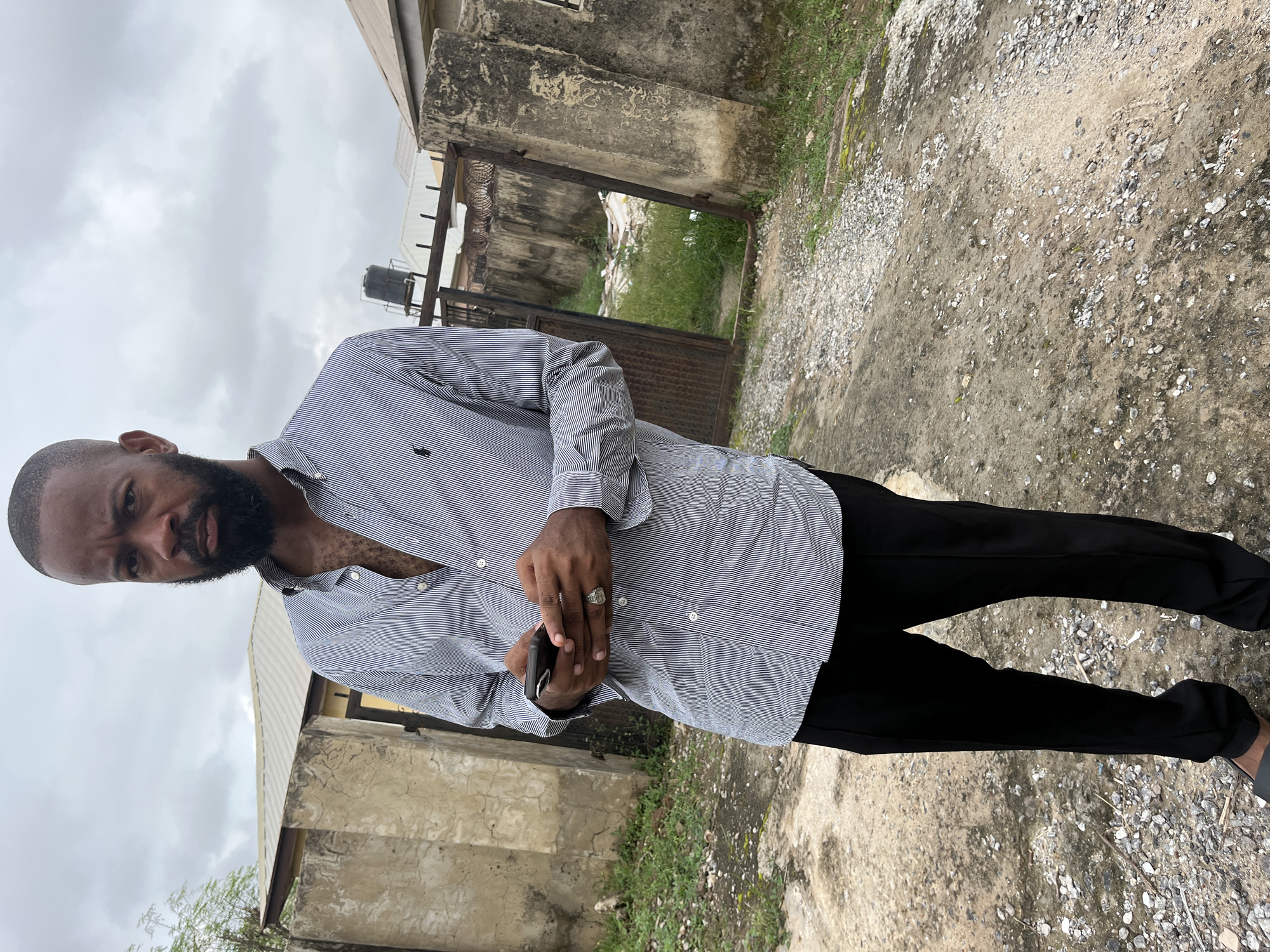 A man stands outside under a cloudy sky, looking directly at the camera with a serious expression. He has a close-cropped haircut and a full beard. He is wearing a light blue and white striped button-down shirt, unbuttoned at the collar, and black pants. He is holding a dark-colored smartphone in his hands, and a silver ring is visible on his right ring finger. The background appears to be an urban or semi-urban environment, possibly in the Gwarinpa district of Abuja, Nigeria. A rough, unpaved path is in the foreground, with scattered gravel and some patches of dry grass. To the left of the man, there is a rough concrete wall with some vegetation growing around it. To the right, there is a rusty metal gate and a concrete wall, partially obscured by foliage. A water tank on a rooftop is visible in the distance. The overall impression is of an overcast day in a developing area.