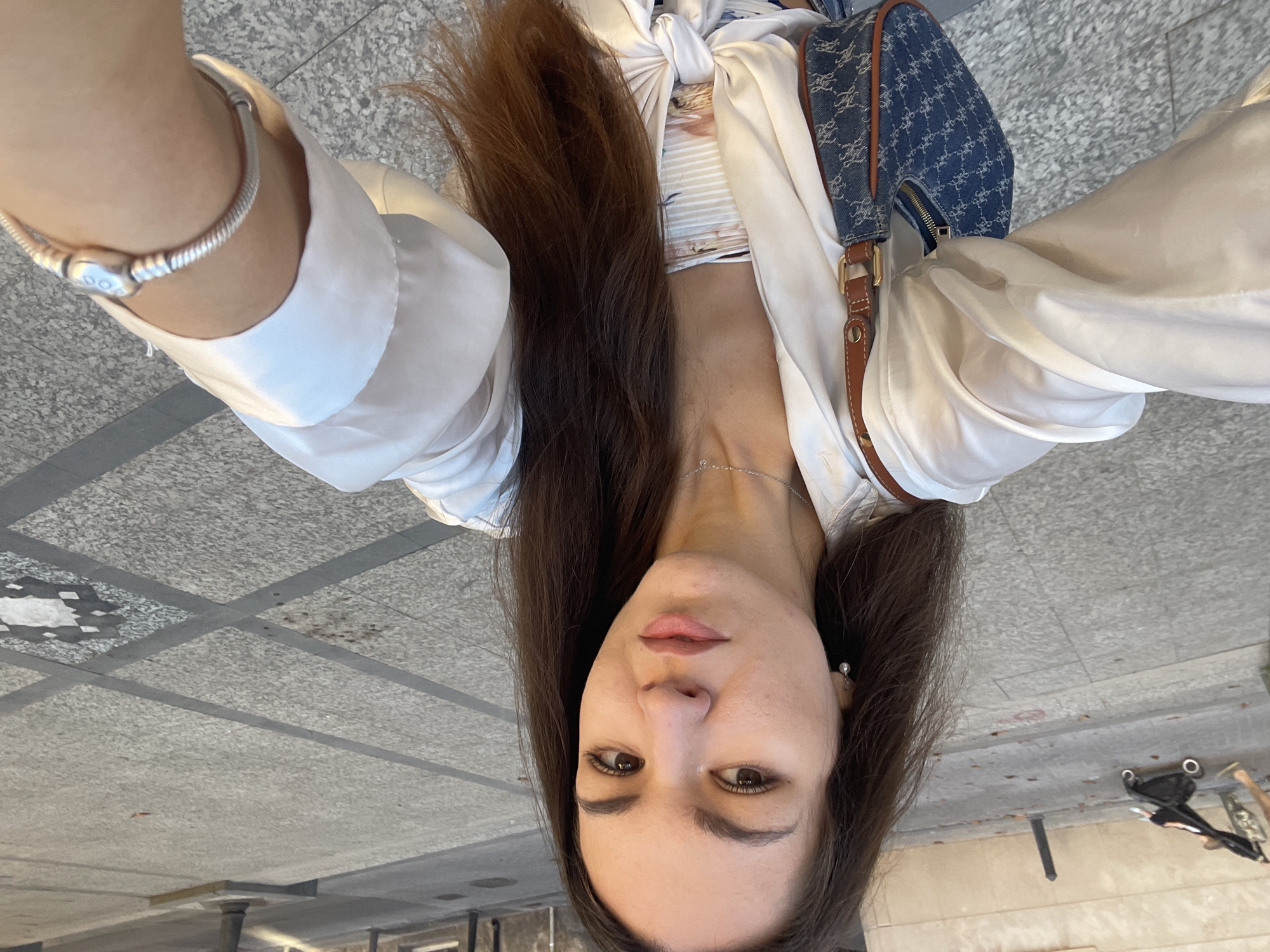 A woman is taking a selfie in what appears to be an urban outdoor location, likely València, Spain, based on visual cues and architectural styles often found in the city. The ground is paved with light-colored stone tiles arranged in a square pattern, with darker gray strips forming a grid. The woman is facing the camera with her head tilted. She has long, brown hair, and she is wearing a white, loose-fitting top or blouse over a patterned shirt. She is also wearing a silver bracelet and a necklace. A blue patterned bag with leather detailing is visible on her shoulder. The background includes parts of street furniture and potentially a building facade.