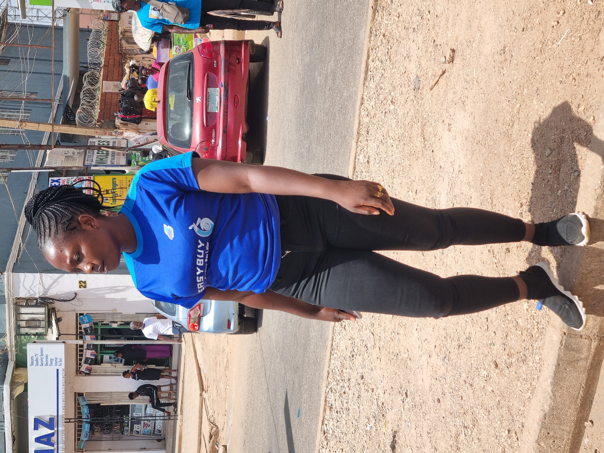 A full-body shot shows a woman wearing a blue EasyBuy T-shirt, black leggings, and black sneakers standing on a sandy and partially paved area. She has dark hair braided into cornrows. She is positioned near a road with parked cars; a red Peugeot is visible. Behind her, there is a building with a sign labeled "AZ" that lists services like "Telecoms," "Satellite TV," "Security Systems," and "Electronics." A second sign, "Tasty Fingers," is present. The text "Jos, Plateau" is visible on the number plate of the red Peugeot. Shops with people inside are visible in the background. The scene suggests a bustling urban environment in Jos, Nigeria.