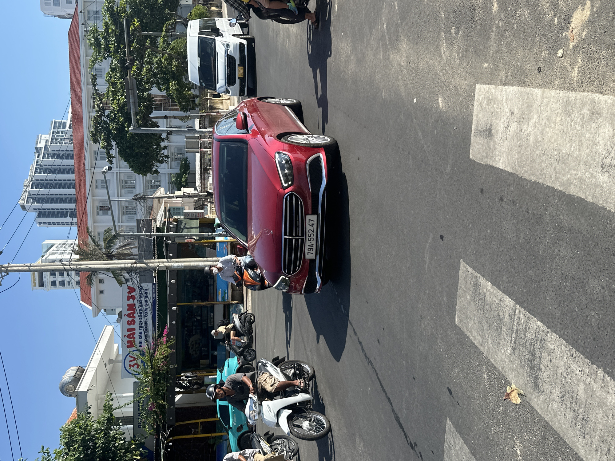The image shows a street scene in Bac Nha Trang Ward, Vietnam. A red Mercedes-Benz car with license plate "79A-552.47" is prominently featured in the foreground. Several motorcycles are positioned around the car, some with riders. A white van is parked in the background. In the background, there's a building with the sign "3V HẢI SẢN 3V," advertising seafood. There are also various buildings, trees, and utility poles. A pedestrian crossing is visible on the road. The scene is taken during the daytime under bright sunlight.