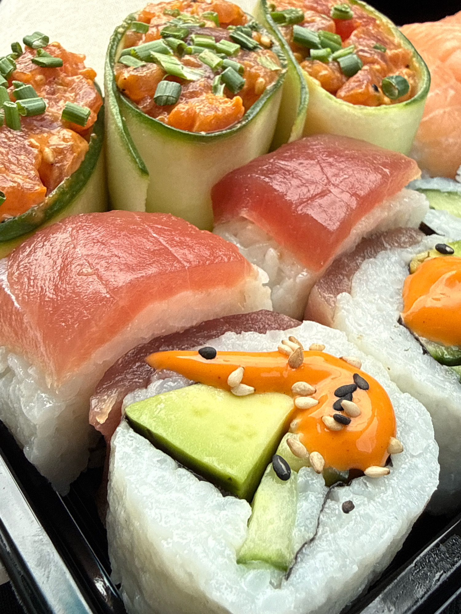 A close-up, overhead shot showcases a selection of sushi. The sushi pieces are arranged in a black plastic container. In the foreground, a sushi roll is visible with white rice, green avocado, and a drizzle of orange sauce topped with sesame seeds and possibly roe. To the left and slightly behind, cucumber rolls filled with what appears to be diced salmon and topped with chopped chives are prominently displayed. Behind these, there are pieces of nigiri sushi with bright red tuna or salmon atop white rice. The lighting is bright and even, highlighting the textures and colors of the food. The background is out of focus, appearing to be a neutral, light-colored surface. The image was taken on an unknown date in Herzberg, Germany.