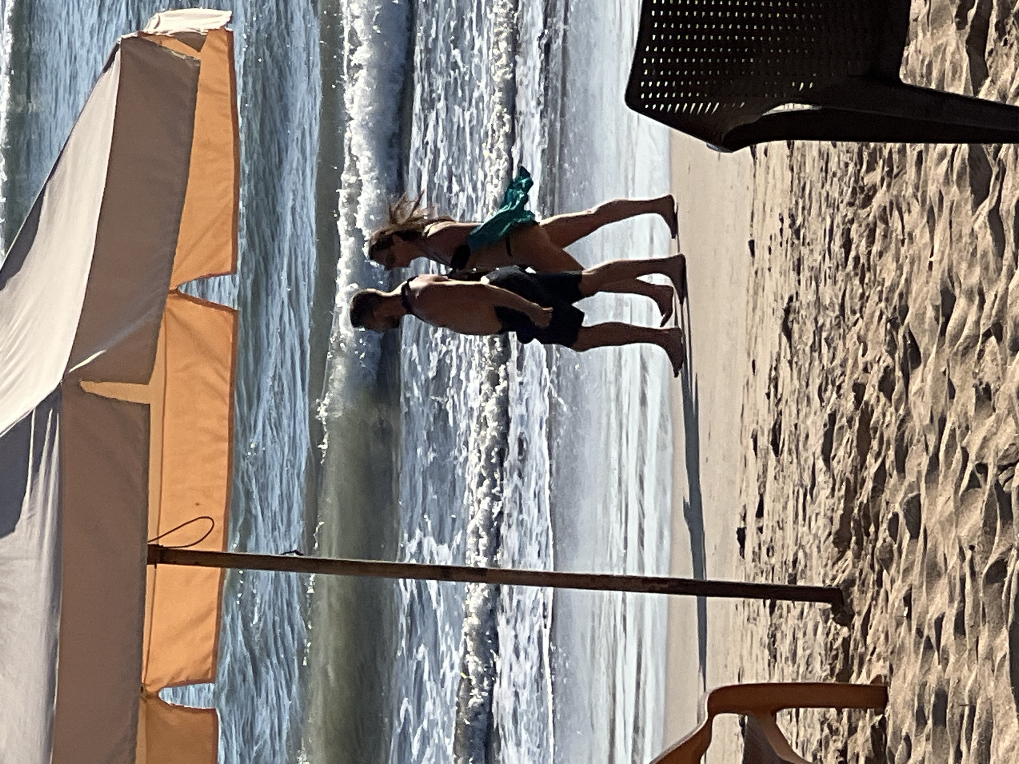 A couple is walking on a sandy beach in Kuta, Indonesia, near the ocean. The man is wearing dark shorts and is shirtless, while the woman is wearing a bikini and carrying a turquoise cloth. They are walking from left to right, with the waves of the ocean behind them. In the foreground, there is an orange beach chair to the left and a dark chair to the right. A beach umbrella is positioned above the couple, with its pole visible. The sand is light brown and textured with footprints and small stones. The ocean water is blue with white waves breaking. The scene appears to be a bright, sunny day.