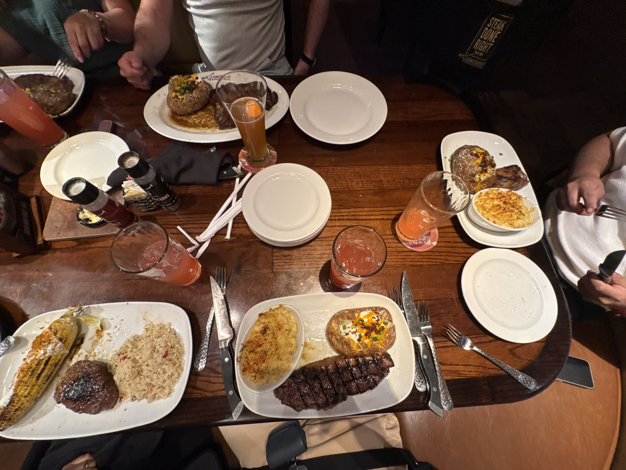 The image captures a dining scene at what appears to be a LongHorn Steakhouse, confirmed by visible text. A dark wooden table is laden with multiple plates of steakhouse fare, suggesting an evening meal due to the warm, dim lighting. Several individuals are partially visible around the table, their hands holding forks and knives as they partake in their meals.
Each diner has a substantial plate, primarily featuring various cuts of grilled steak. Accompaniments include loaded baked potatoes topped with cheese, bacon bits, and chives; creamy mac and cheese in oval ramekins; seasoned rice; and grilled corn on the cob. Beverages include multiple glasses of a pink, possibly fruit-flavored, drink and one tall glass of an amber-colored beer or ale.
White rectangular and round plates, forks, knives with dark handles, and black salt and peppercorn grinders are neatly arranged on the table. Visible text on a bag or menu item clearly states "STEAK DONE RIGHT" and "LongHorn Steakhouse", with smaller labels on the grinders indicating "SALT" and "PEPPERCORNS". The scene depicts a casual American steakhouse in Schaumburg, United States of America, with an atmosphere of people enjoying a hearty meal together.