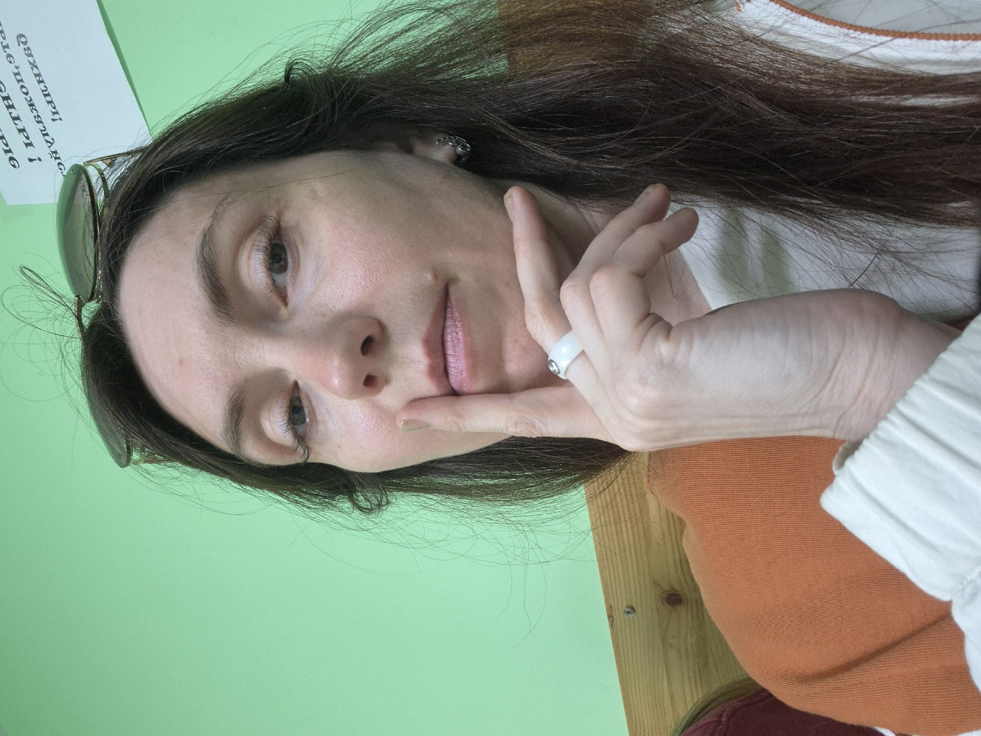 A woman is posing for a selfie in an indoor setting with a light green wall and a wooden desk visible in the lower right corner. She is wearing a white and orange top, with dark brown hair. She has sunglasses perched on top of her head, and is wearing a white ring on her left hand. She is resting her chin on her index finger and looking slightly down at the camera with a gentle smile. There is a sign on the wall with illegible text. The location is Unknown, Unknown.