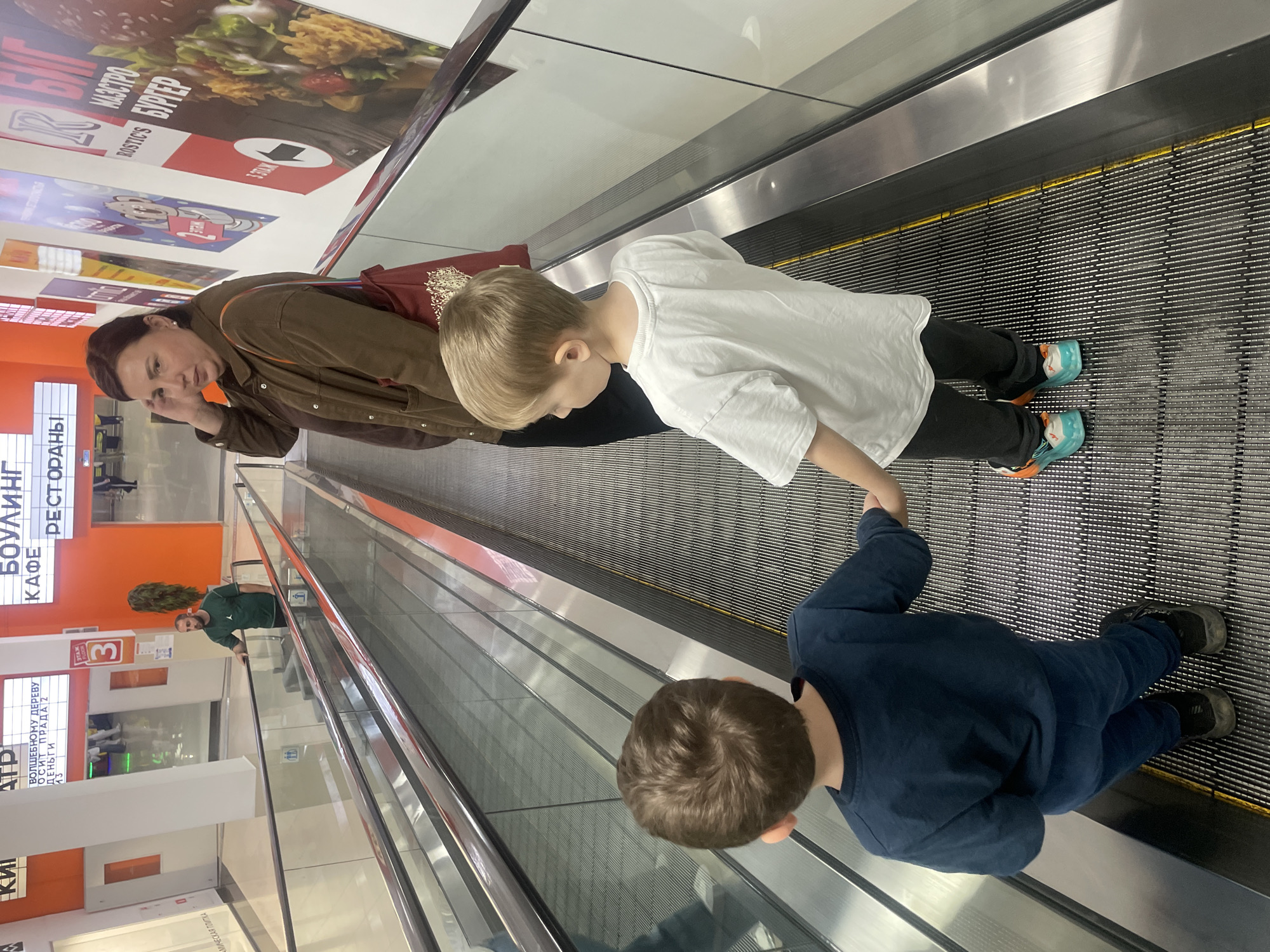 An adult woman and two young boys are riding an escalator in a commercial building in the city of Sochi, Russia. The woman is talking on a mobile phone. One of the boys is standing, while the other is sitting on the escalator holding the other child's hand. In the background is a signage for "Bowling" and "Restaurants" in Russian, as well as advertising for "Rostic's" burgers. Another man in a green shirt is also visible on the escalator.