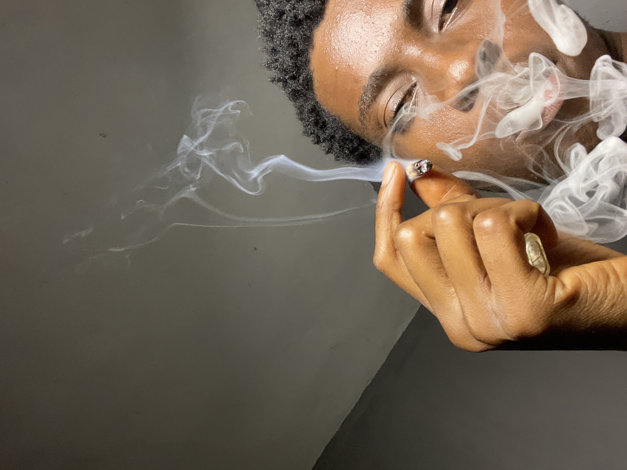 This media file features a close-up shot of a person's face and hand, with wisps of smoke prominently visible. The person is holding a lit cigarette between their thumb and index finger, exhaling smoke that billows around their face. Their eyes are closed, and their expression appears relaxed. The background is a plain, dark gray surface, suggesting an indoor setting. There are no visible text elements in the image. The scene is lit in a way that highlights the smoke and the contours of the person's face and hand. The overall mood is intimate and contemplative.