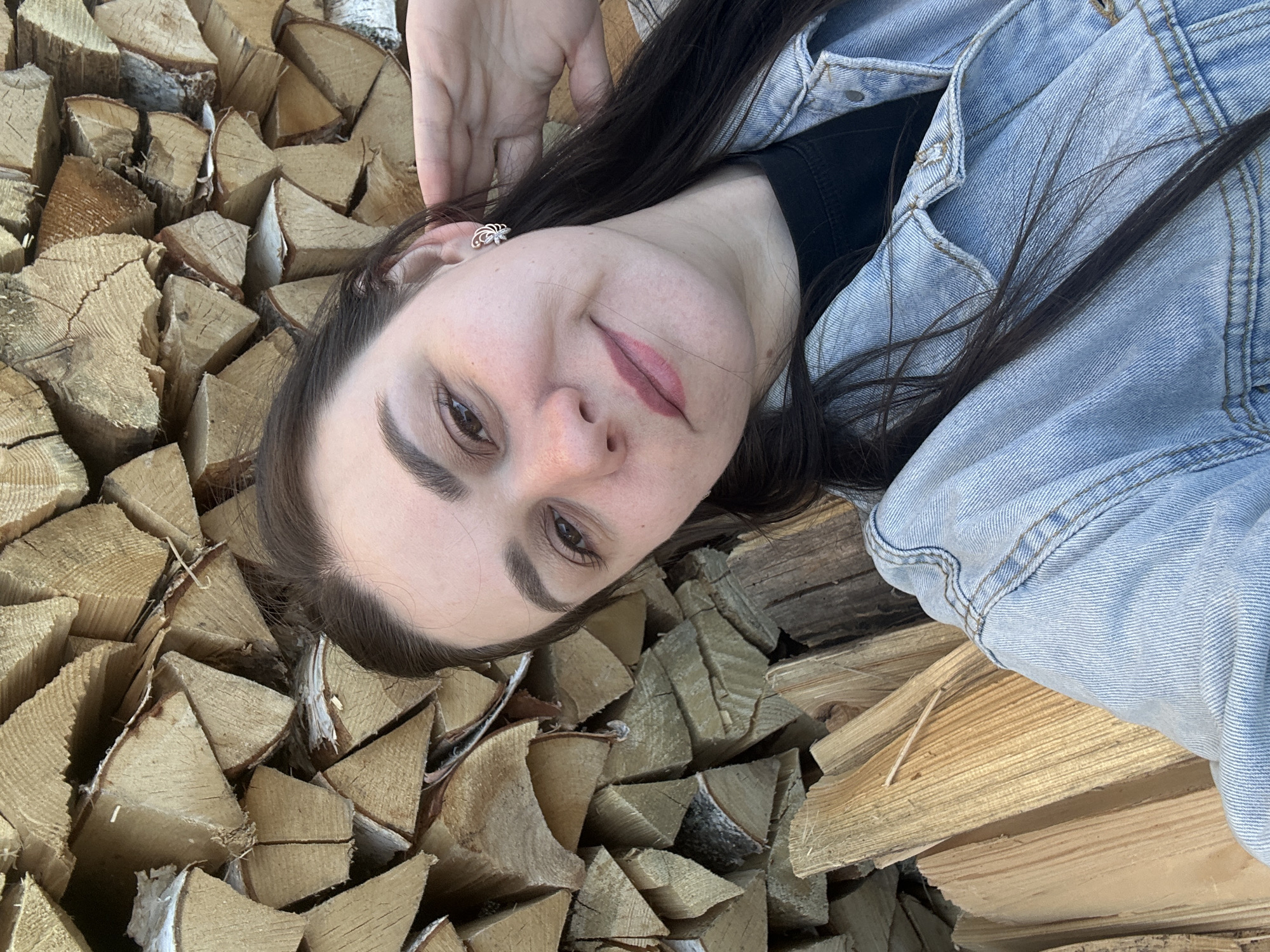 A woman is lying on a pile of firewood. She has long dark hair and is wearing a denim jacket over a black shirt. She is looking at the camera. The firewood is mostly birch and is stacked haphazardly. It is not possible to verify the location, city of Chaikovsky, Russia, from the visual information.