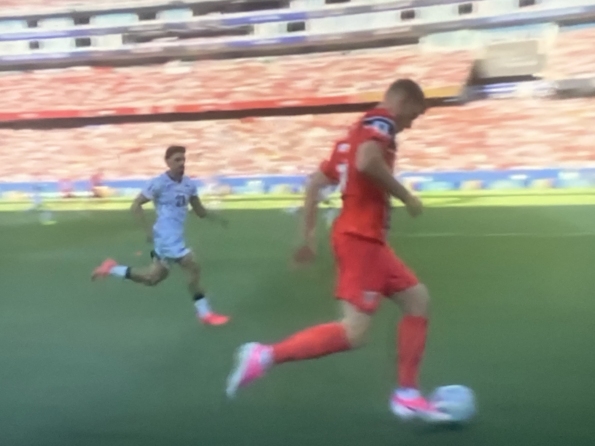 Two male soccer players are on a green field in a stadium. One player, wearing a red jersey and shorts with red socks, is kicking a white soccer ball. The other player, wearing a light blue jersey with a number 21 on it, shorts, and white socks, is running away from the first player. In the background, there are stadium seats filled with people, appearing to be a large crowd. The overall image is somewhat blurry, suggesting it might be a still frame from a video. The scene depicts a moment of action during a soccer match.
