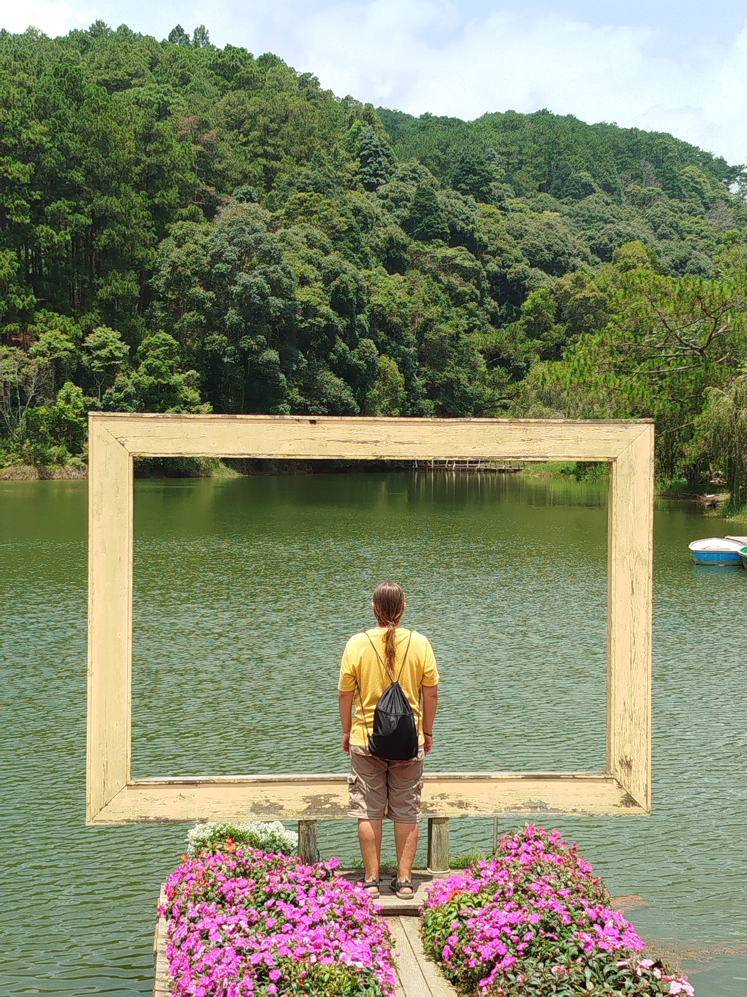 The image depicts a person standing on a wooden platform at Cam Ly Lake in Da Lat, Vietnam, facing a large, weathered, yellow picture frame. The frame is positioned in front of the lake, framing a view of the lush green forest that covers the hills beyond. The person, viewed from behind, wears a yellow t-shirt, khaki shorts, and a black backpack. They are standing with their feet slightly apart, looking out at the scenery. In the foreground, on either side of the platform, are vibrant displays of pink flowers, with some white flowers interspersed. The lake's surface is rippled, reflecting the green of the surrounding foliage and the sky. In the distance, across the lake, a wooden pier is visible. To the right of the frame, on the lake, two small boats are docked. The overall scene suggests a serene and picturesque natural environment, likely a tourist attraction.