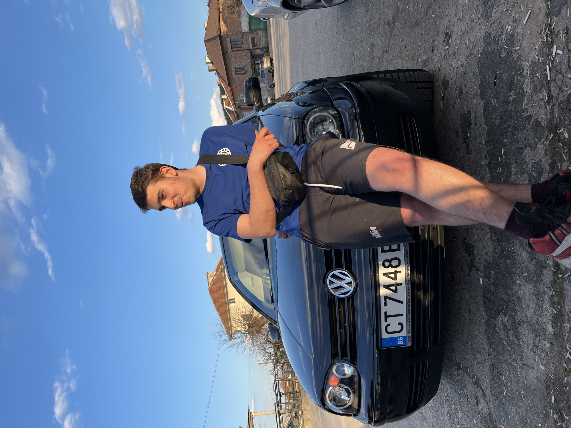 A young man leans against the hood of a black Volkswagen Golf with Bulgarian license plate CT 7448 E. He is wearing a blue t-shirt, black shorts with KTM Racing branding, and athletic shoes. He has a black waist bag slung over his shoulder. The car is parked on an asphalt surface strewn with cigarette butts. In the background, buildings can be seen under a blue sky with scattered clouds. The buildings suggest an urban or suburban environment, indicating the location is likely Karavelovo, Bulgaria, based on the license plate origin.
