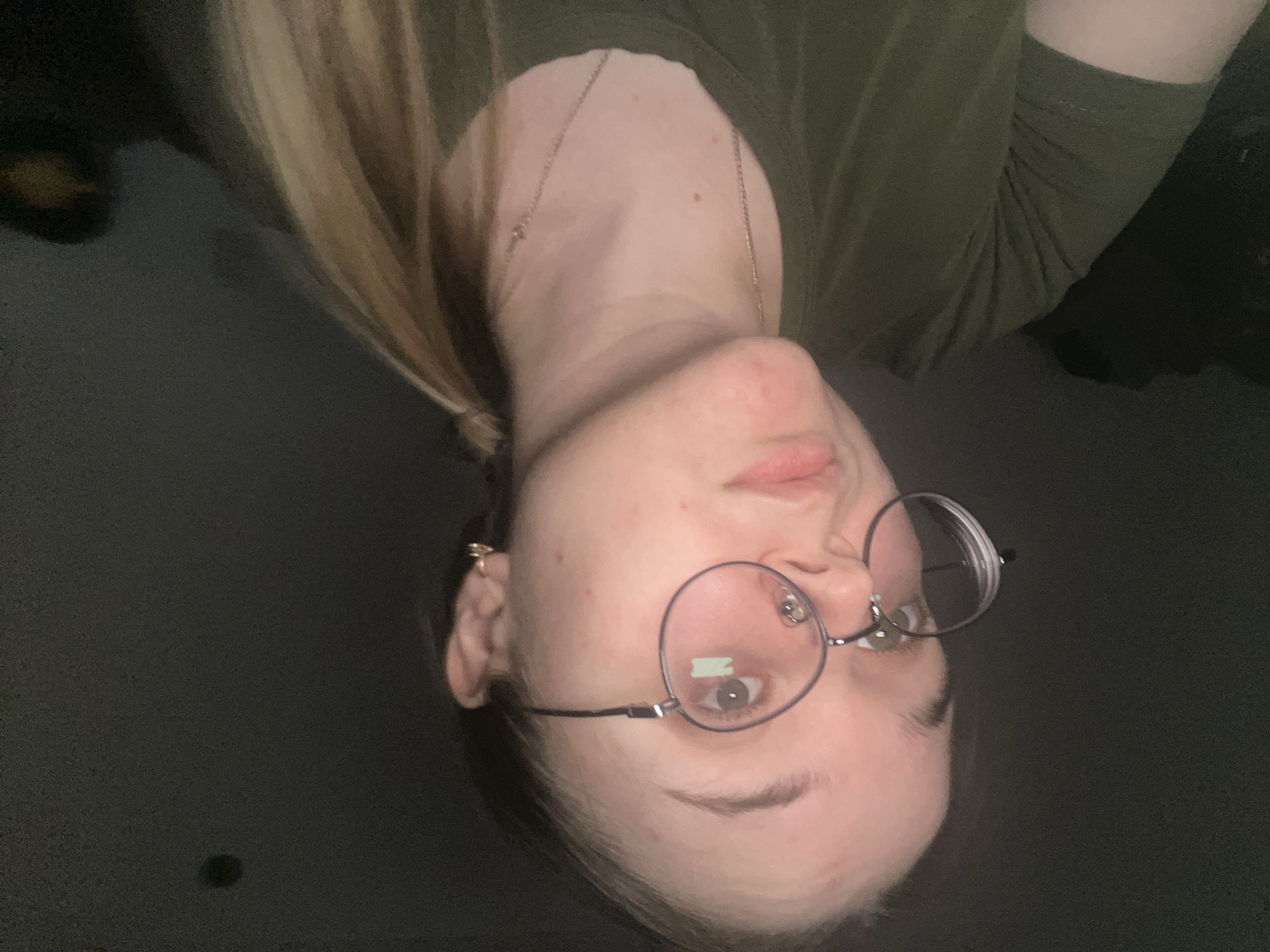 An image taken in Pushchino, Russia, depicts a young adult Caucasian female from the chest up, positioned slightly off-center to the left. She has light-toned skin, light brown or blonde hair tied back in a ponytail visible over her right shoulder, and is wearing round, silver-rimmed eyeglasses. A faint greenish reflection is visible on her left eyeglass lens, just below the pupil. Her eyes are open, and she is looking upwards and slightly to her left, with a neutral expression and a slightly pouting mouth. She wears an olive-green short-sleeved t-shirt, a thin gold-colored necklace with a small pendant, and a small gold-colored hoop earring in her visible left ear. The background is uniformly dark and indistinct, lacking any discernible features or objects, suggesting a low-light environment. Illumination appears to originate from the front, highlighting her face and upper body.